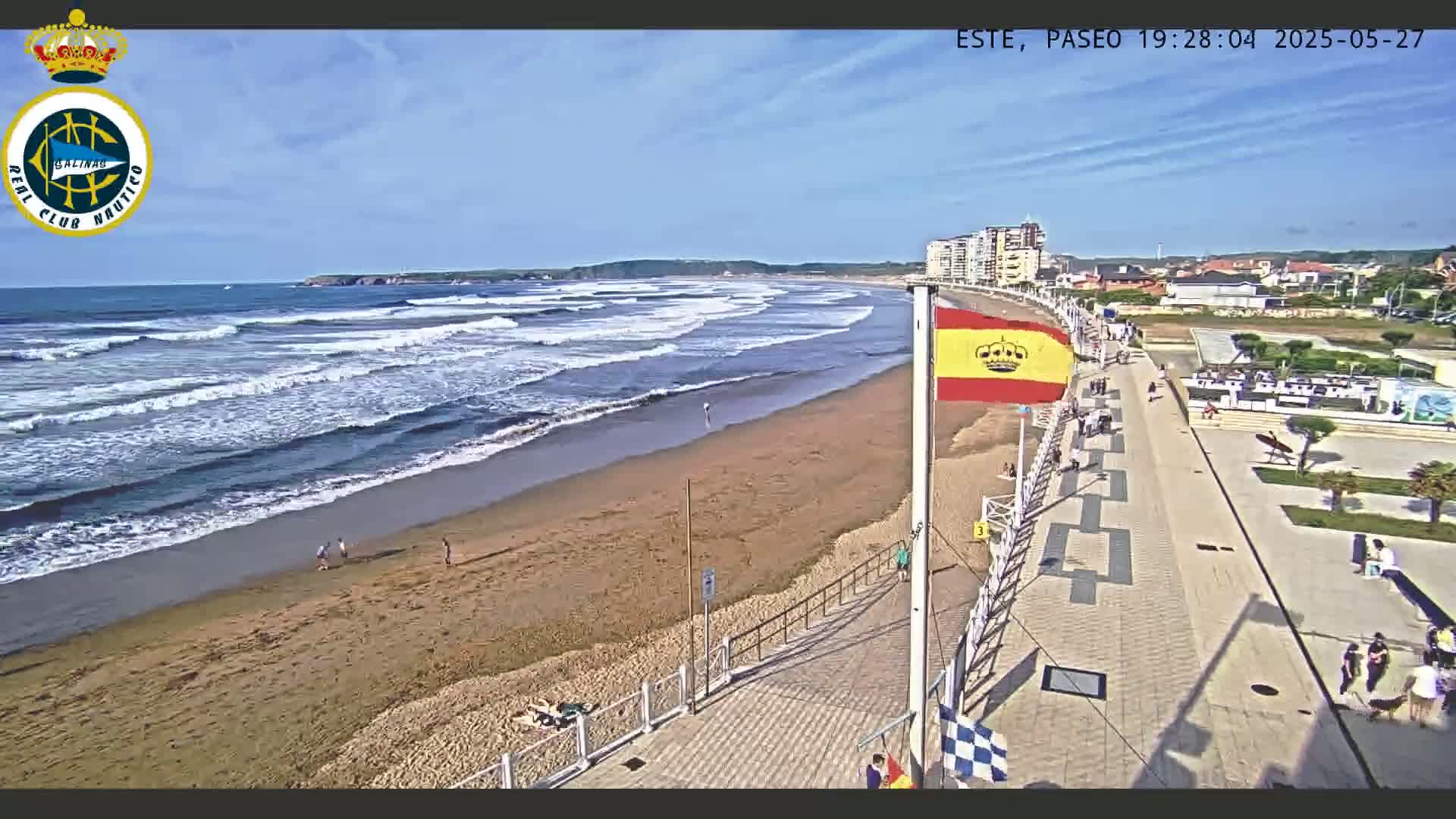 A sandy beach with moderate waves under a mostly sunny sky is bordered by a paved walkway with people walking along it and buildings in the distance.