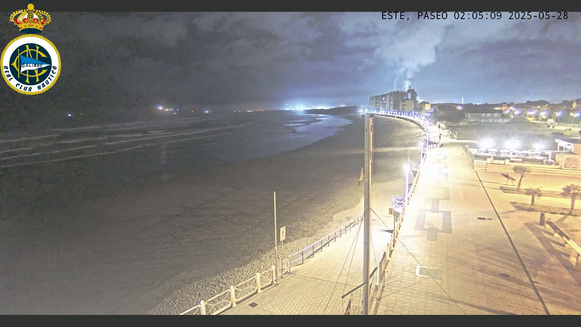 A nighttime view of a mostly empty beach with a boardwalk and buildings in the background under a cloudy sky.