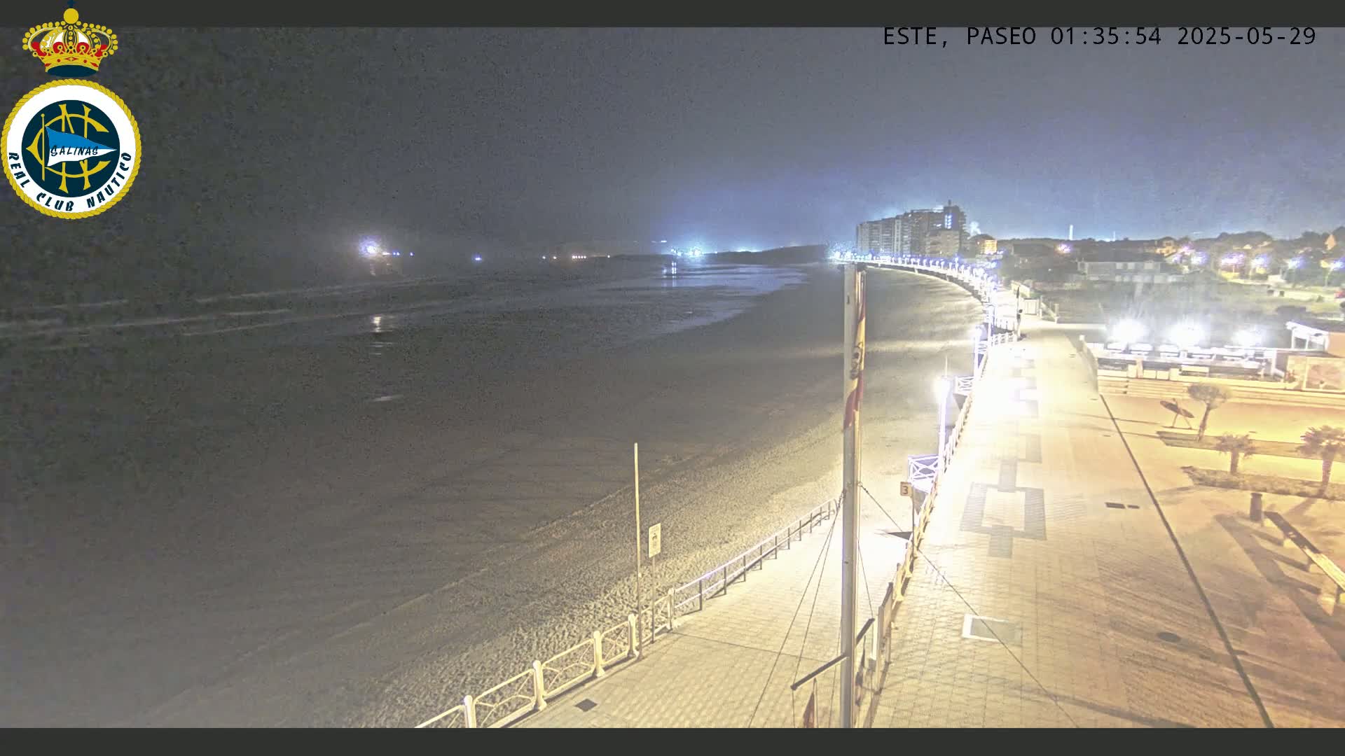 A nighttime view of a mostly empty beach and boardwalk, with buildings and lights visible in the distance under a dark sky.
