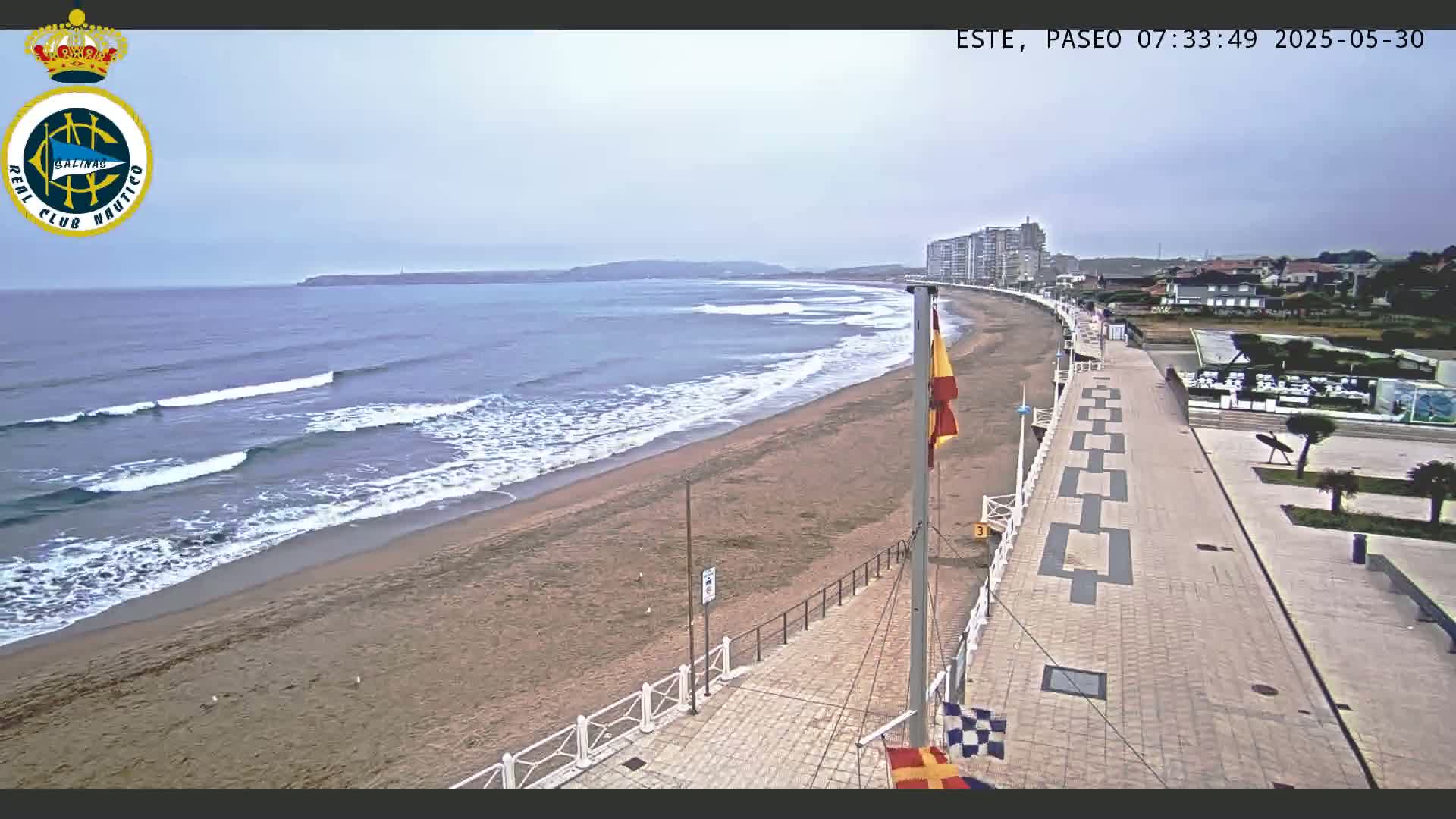 A sandy beach with gentle waves under an overcast sky is bordered by a paved walkway leading to buildings and a grassy area.