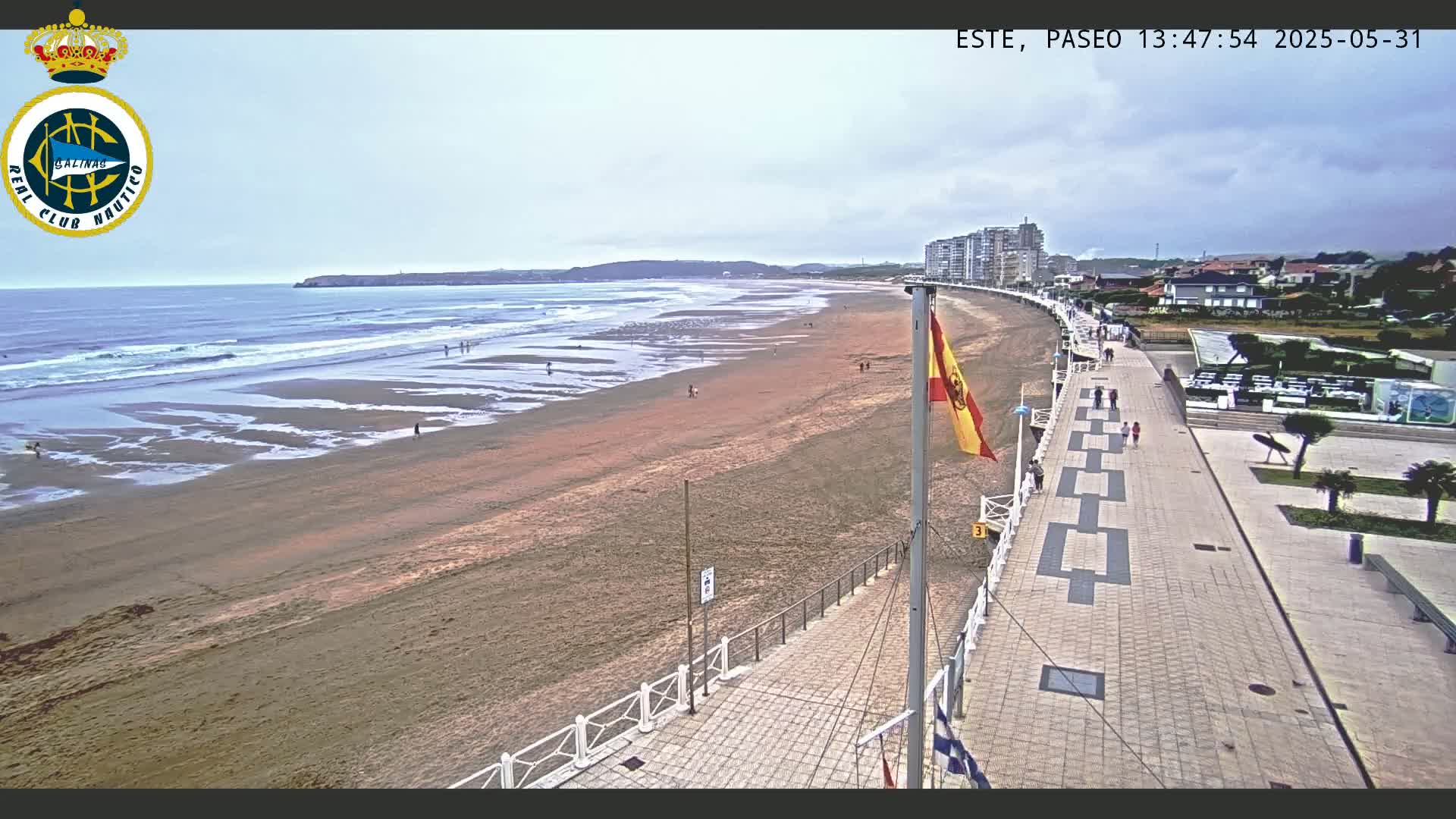 A sandy beach with small waves under an overcast sky is bordered by a paved walkway lined with buildings and a few people strolling.