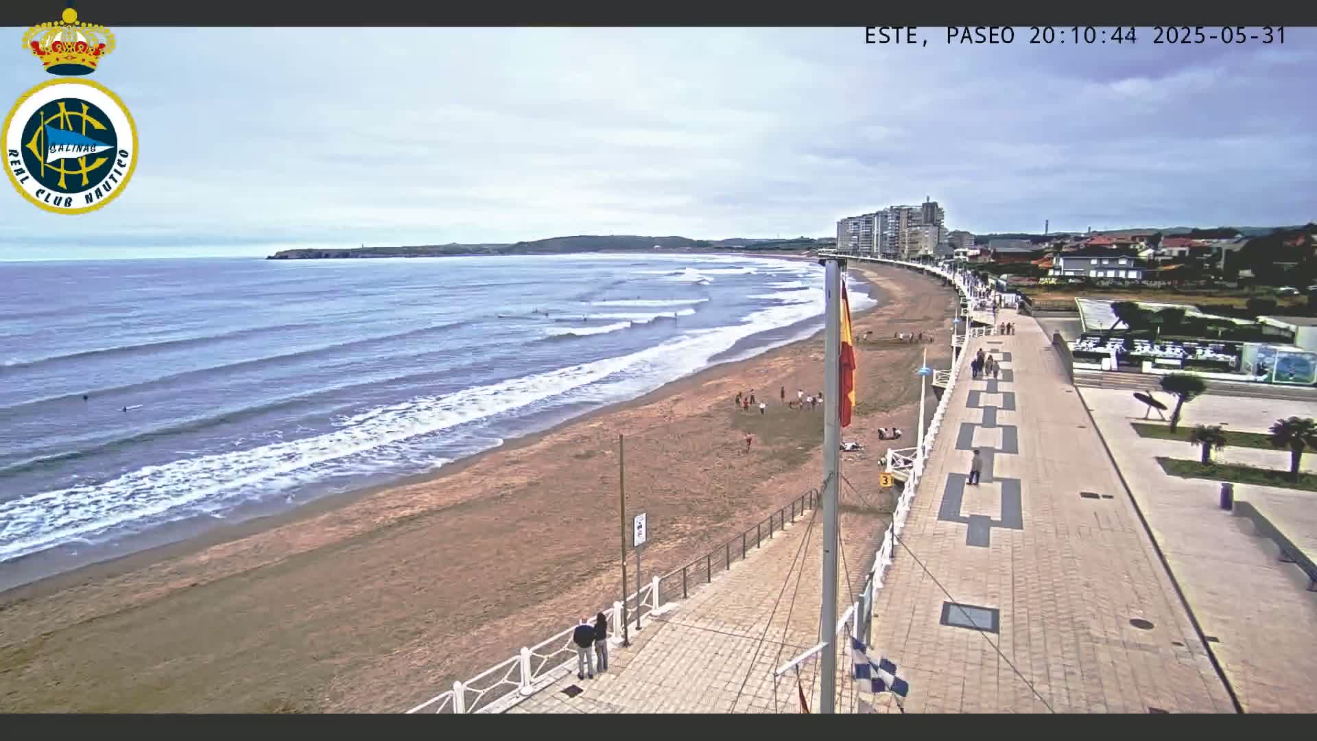 An aerial view shows a sandy beach with gentle waves under a partly cloudy sky, alongside a paved boardwalk with a few people walking, and buildings in the distance.