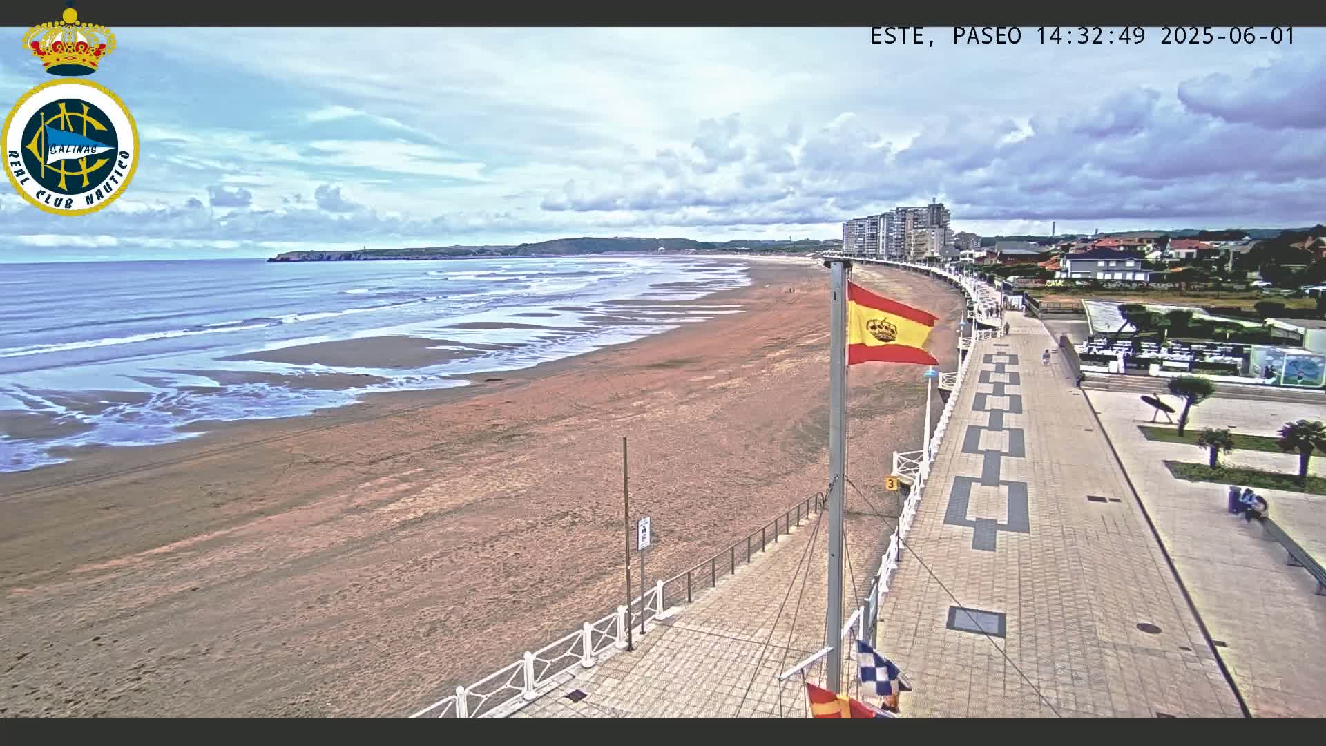 A sandy beach with gentle waves under a partly cloudy sky is seen from a high vantage point, overlooking a paved boardwalk with a Spanish flag and buildings in the distance.