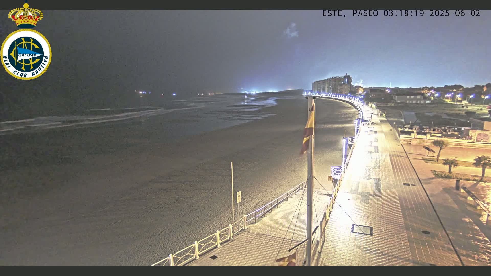 A nighttime view of a beach with a paved walkway, illuminated by streetlights, leading to buildings in the distance under a dark, cloudy sky.