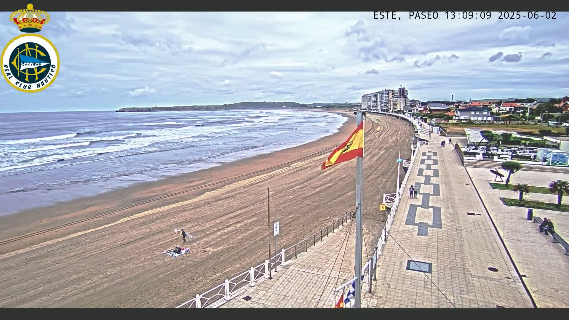 A sandy beach with moderate waves under a partly cloudy sky is bordered by a paved walkway leading to buildings and a plaza.