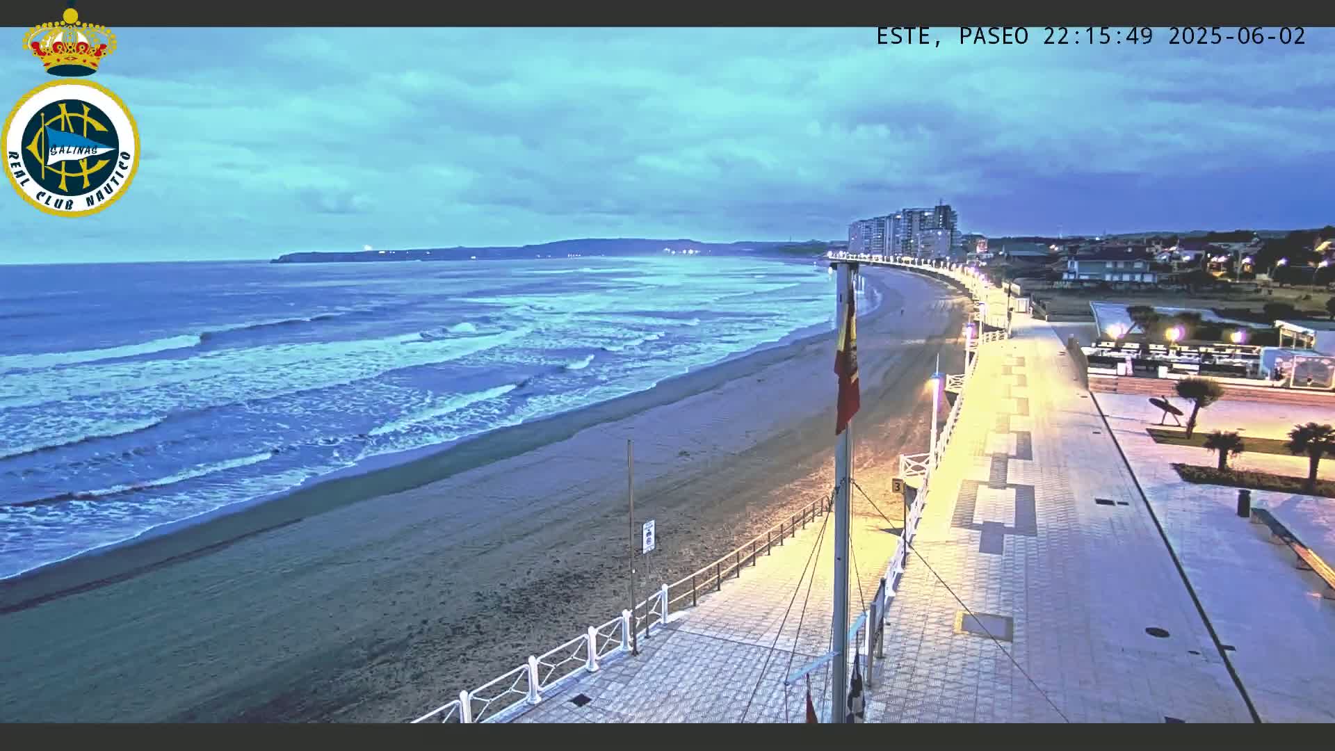 A beach at twilight shows moderate waves lapping a sandy shore, with a paved walkway and buildings visible in the background under a partly cloudy sky.