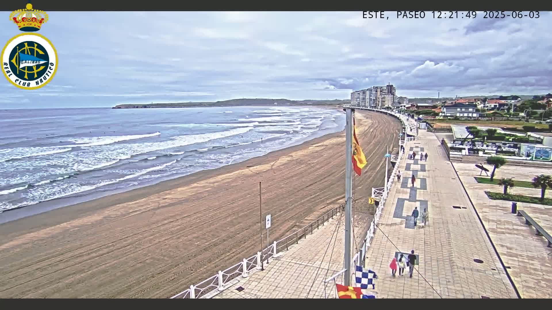 A sandy beach with moderate waves under a partly cloudy sky is viewed from a boardwalk with several people walking along it.