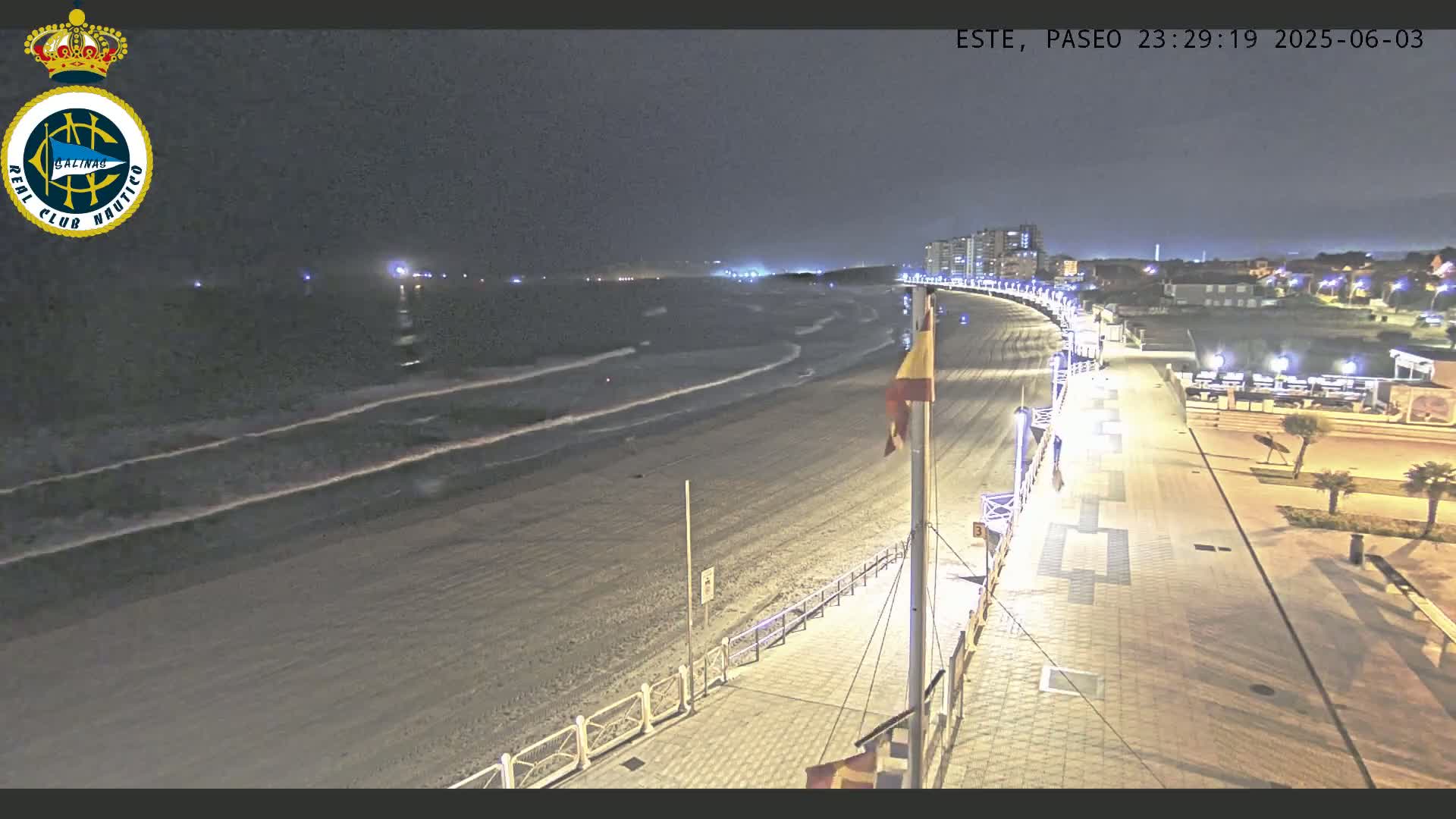 A nighttime view of a beach with gently rolling waves, a paved walkway, and buildings in the distance under a dark sky.