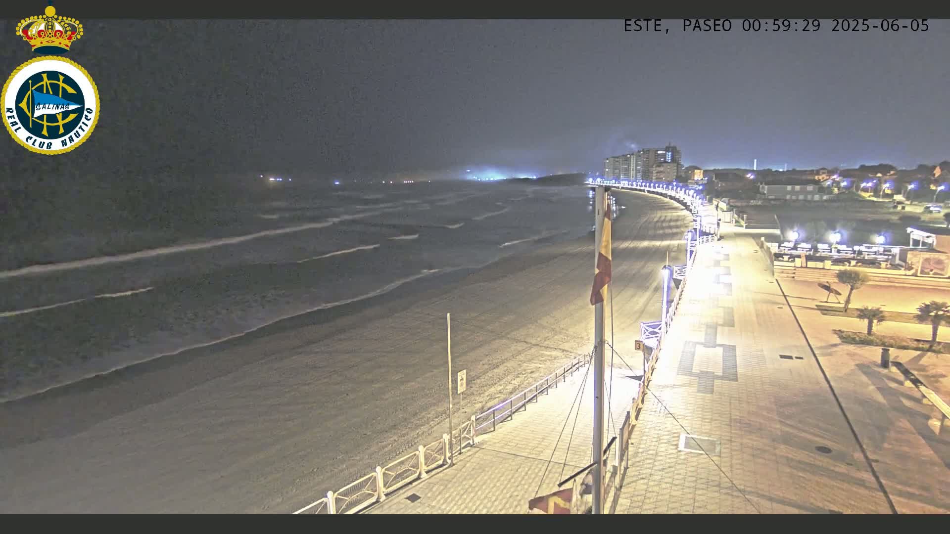 A nighttime view of a beach with moderate waves, a paved boardwalk lined with lights, and buildings in the distance under a dark, clear sky.
