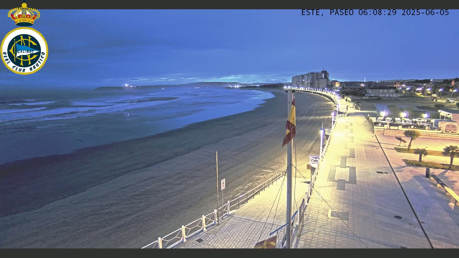 A dark-blue twilight sky overlooks a long, sandy beach with gentle waves, a paved boardwalk with lights, and buildings in the distance.