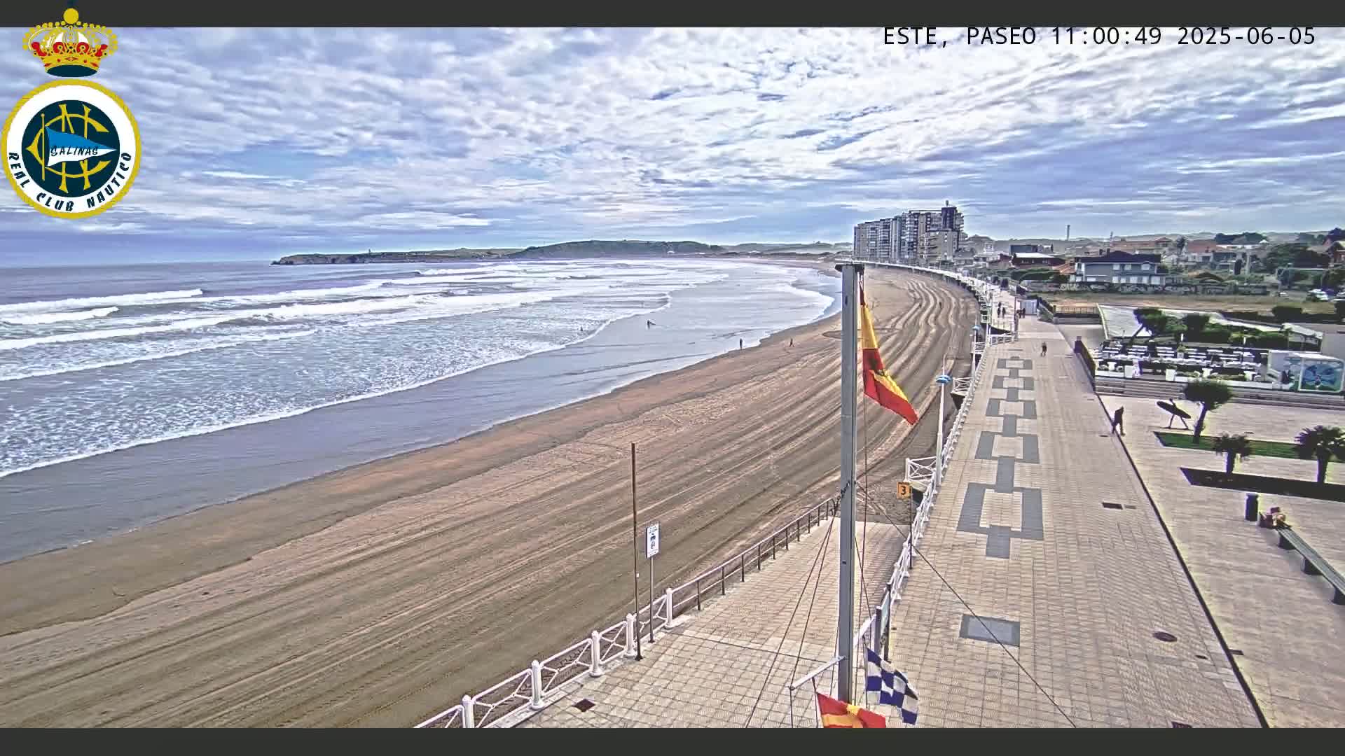 A sandy beach with gentle waves under a partly cloudy sky is bordered by a paved walkway and buildings.