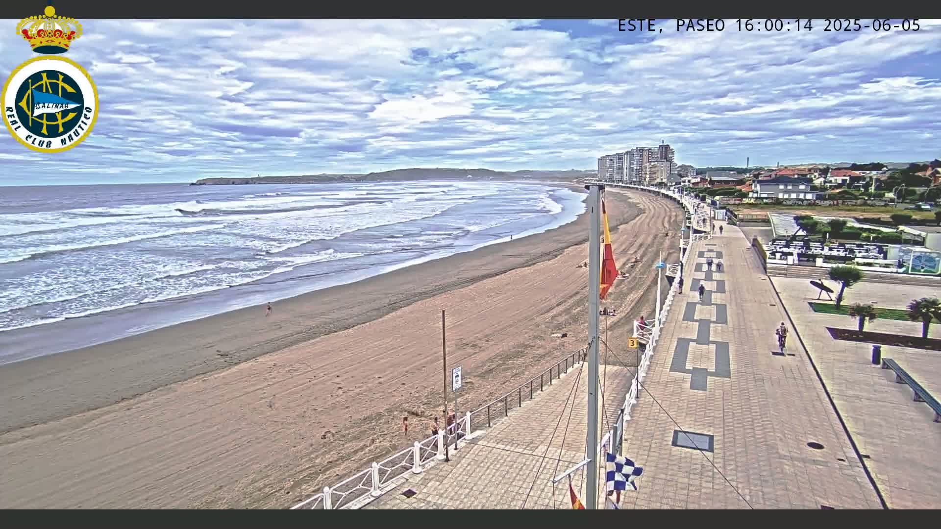 A sandy beach with moderate waves under a partly cloudy sky is viewed from above, showing a paved boardwalk with a few pedestrians and a beachfront area with buildings in the background.