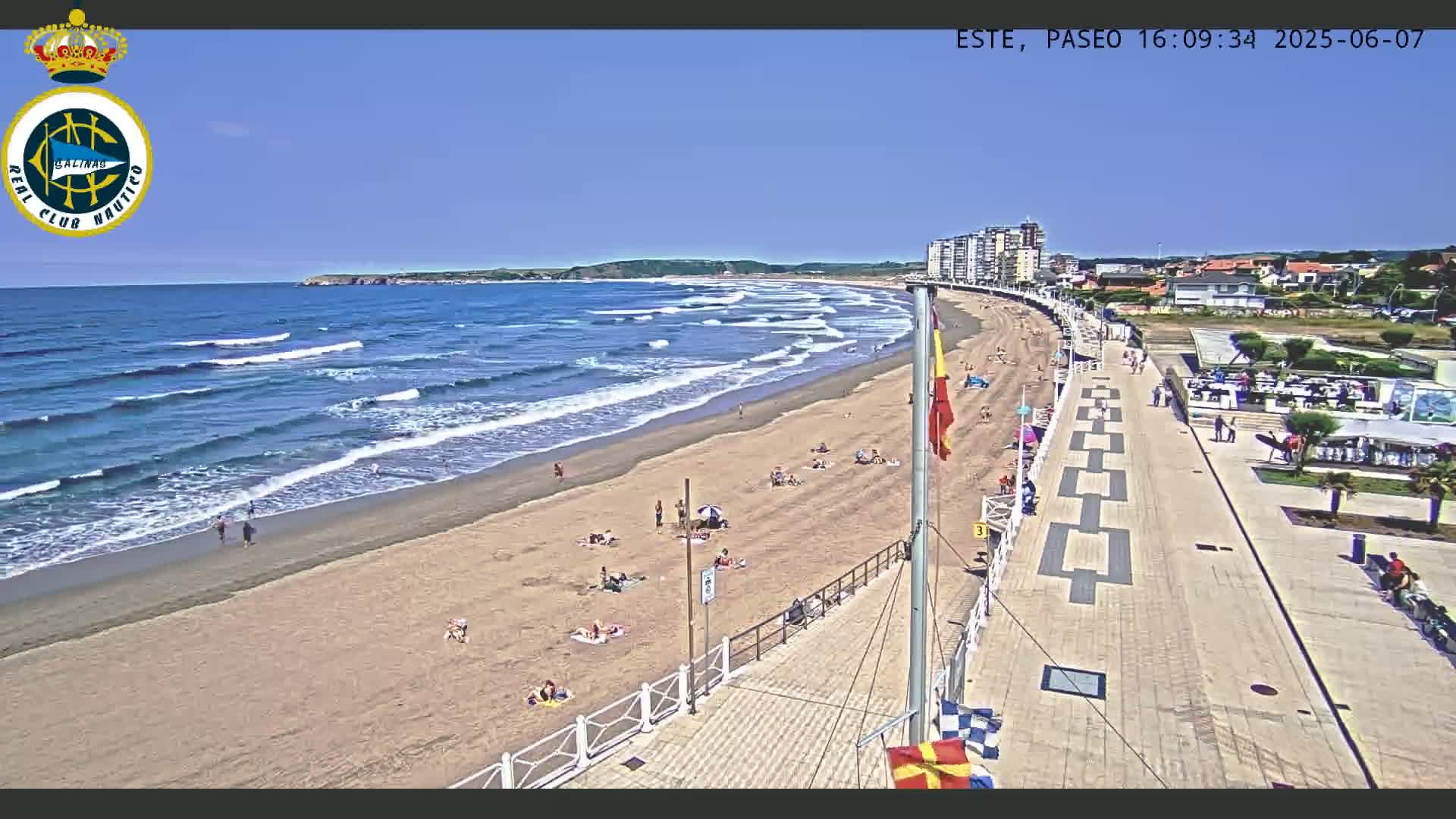 A sandy beach with moderate waves under a clear blue sky is lined with sunbathers and a paved boardwalk with buildings in the background.