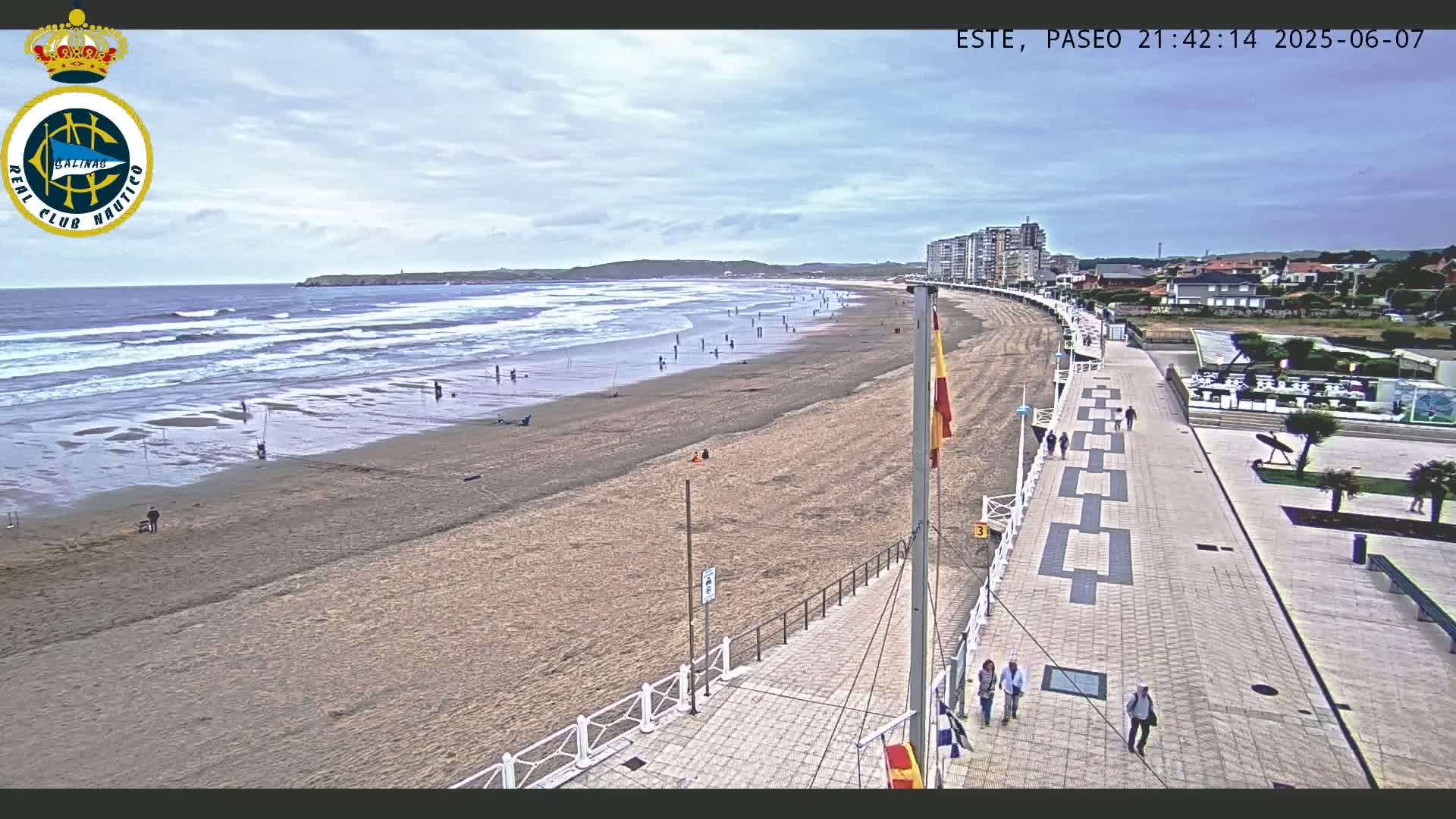 A sandy beach with gentle waves under a partly cloudy sky is bordered by a paved walkway lined with people, leading to buildings along the coast.