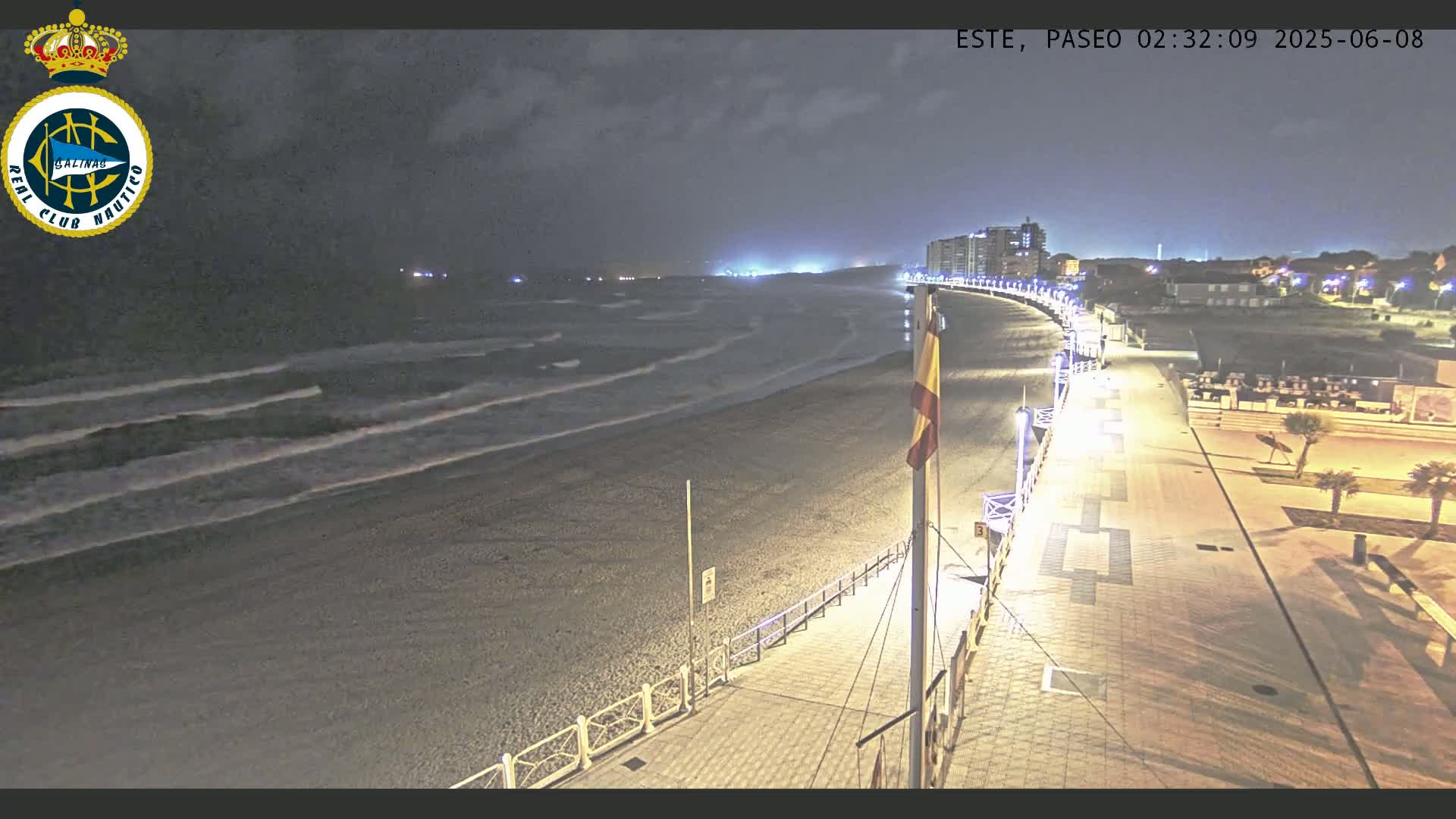 A nighttime view of a beach with gently rolling waves under a dark sky, showing a paved walkway and buildings in the distance.