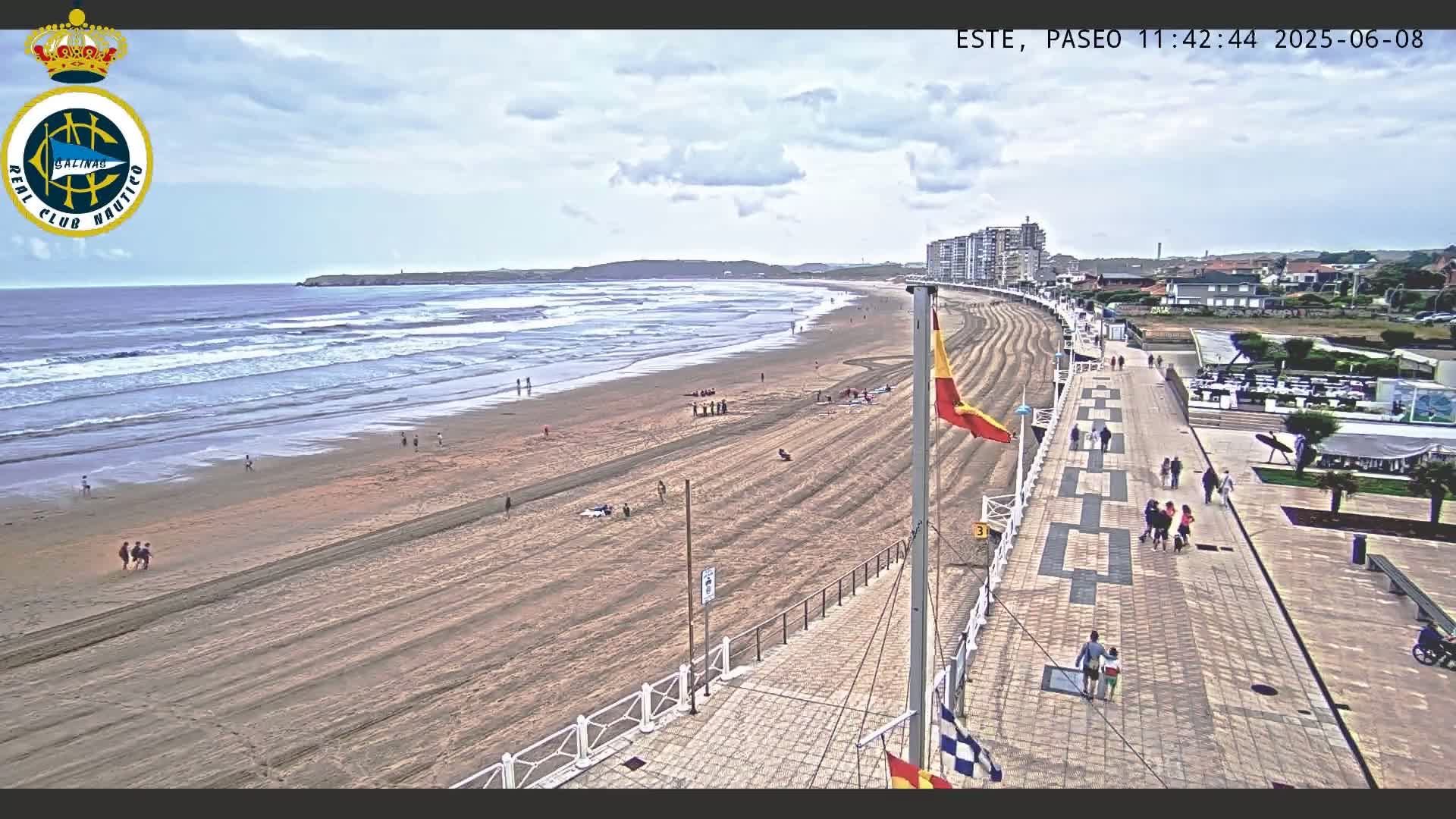 A wide shot of a sandy beach with gentle waves under a partly cloudy sky, showing people strolling along a paved boardwalk that runs parallel to the shore, with buildings visible in the distance.