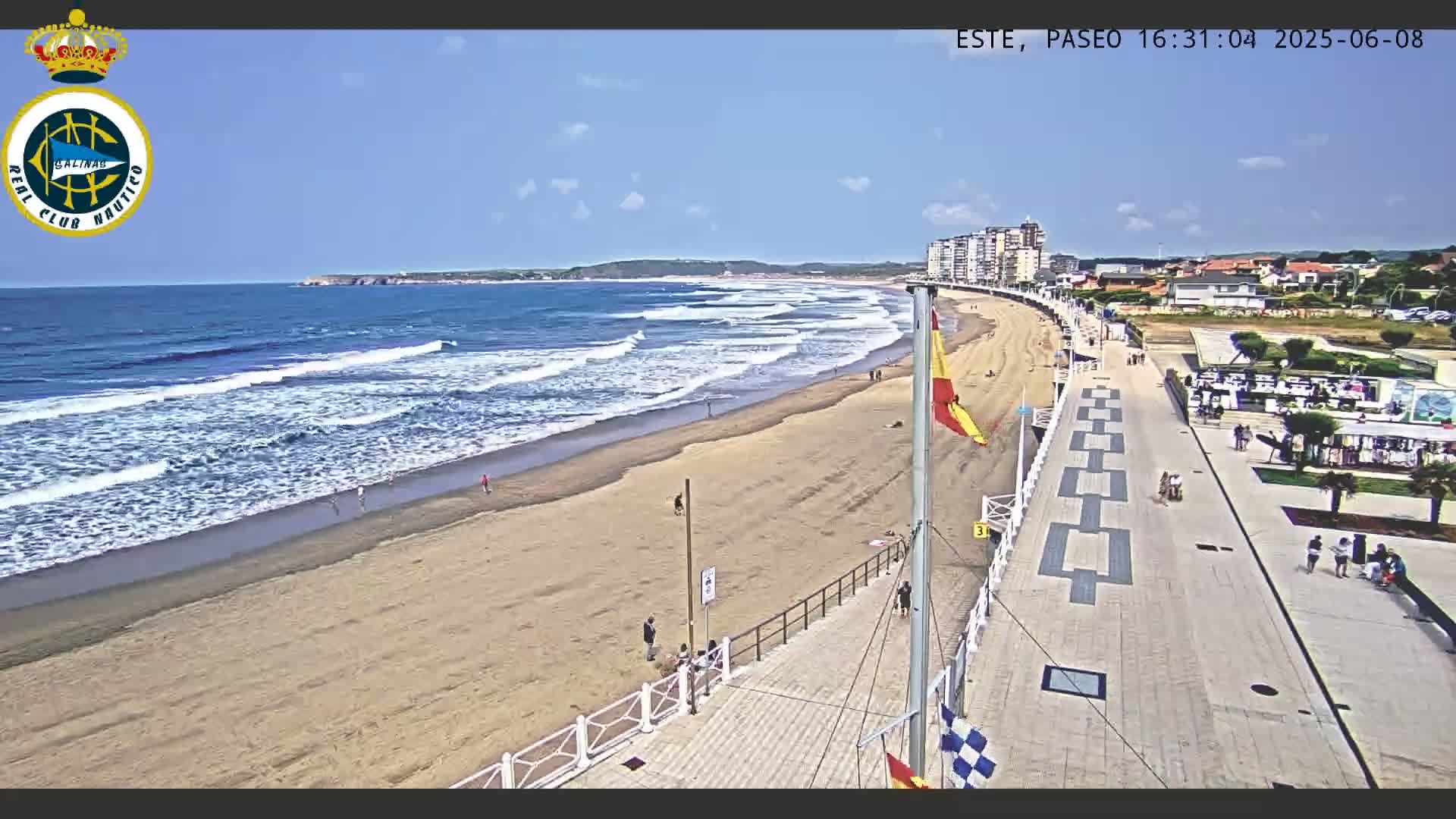 A sandy beach with gentle waves under a clear blue sky is bordered by a paved promenade and buildings.