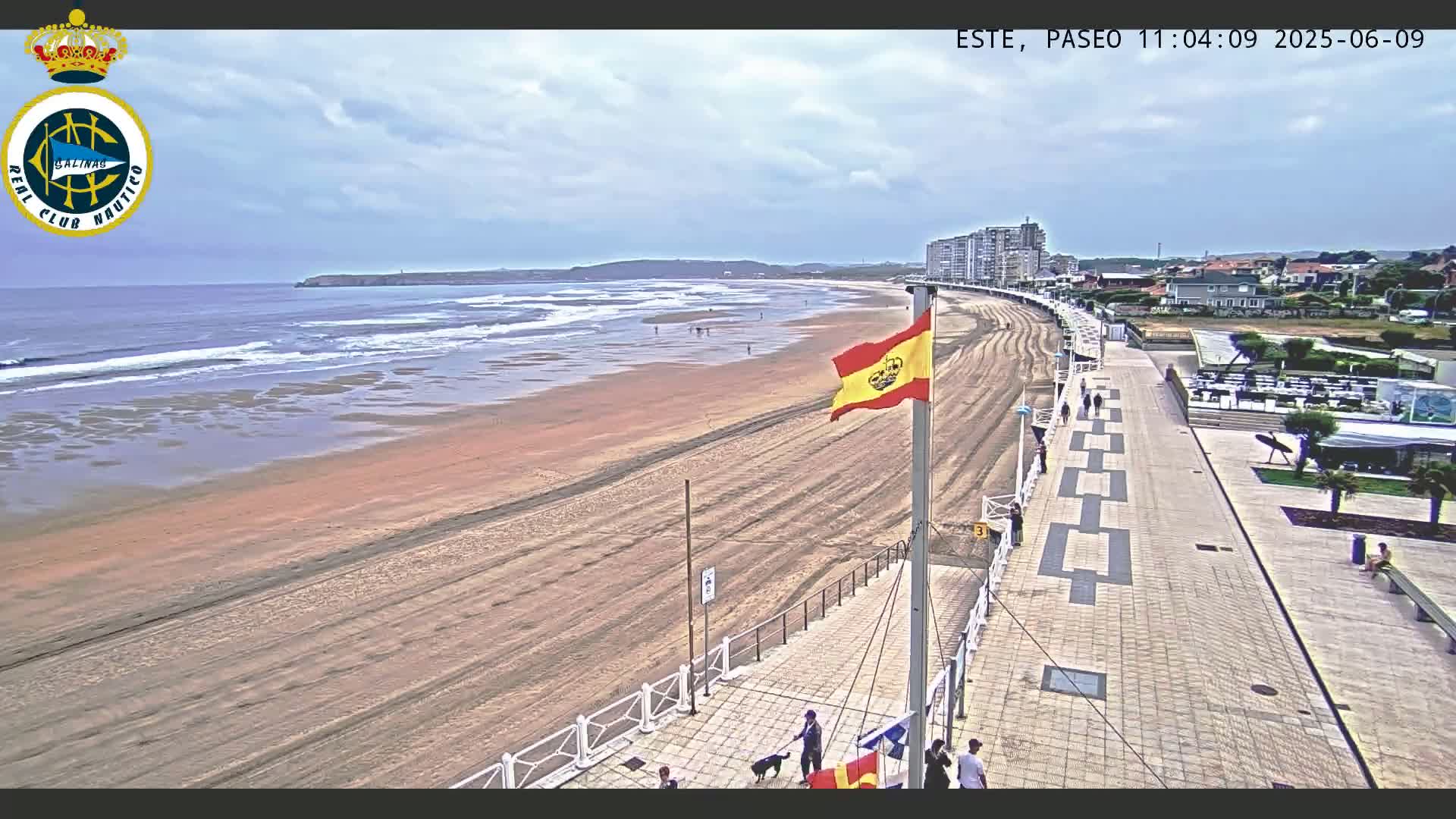 A sandy beach with gentle waves under a partly cloudy sky is bordered by a paved walkway and buildings, with a Spanish flag flying near the shore.
