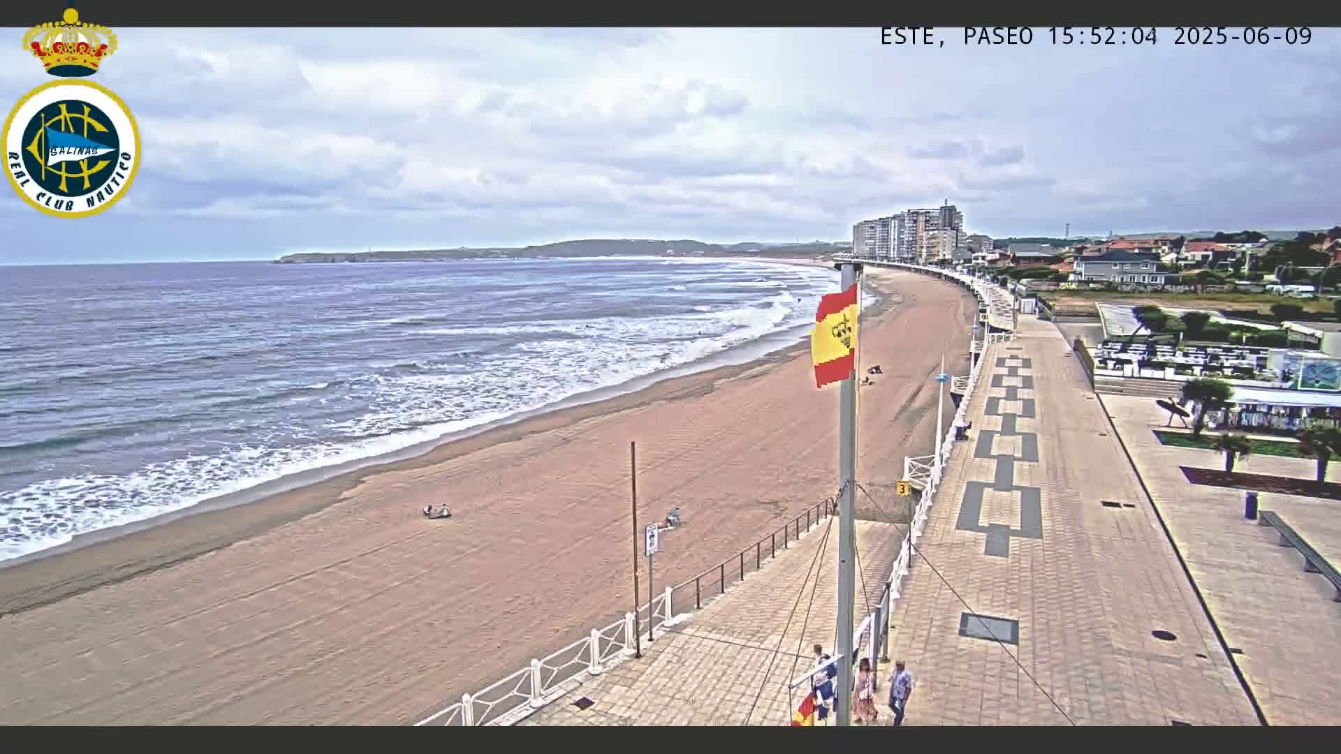 An overhead view shows a sandy beach with gentle waves under a partly cloudy sky, a paved boardwalk with a few people walking on it, and buildings in the distance.