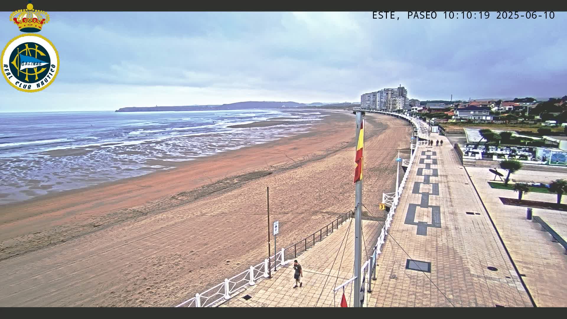 A wide sandy beach stretches out to a gently-lapping ocean under an overcast sky, with a paved boardwalk and buildings visible in the distance.