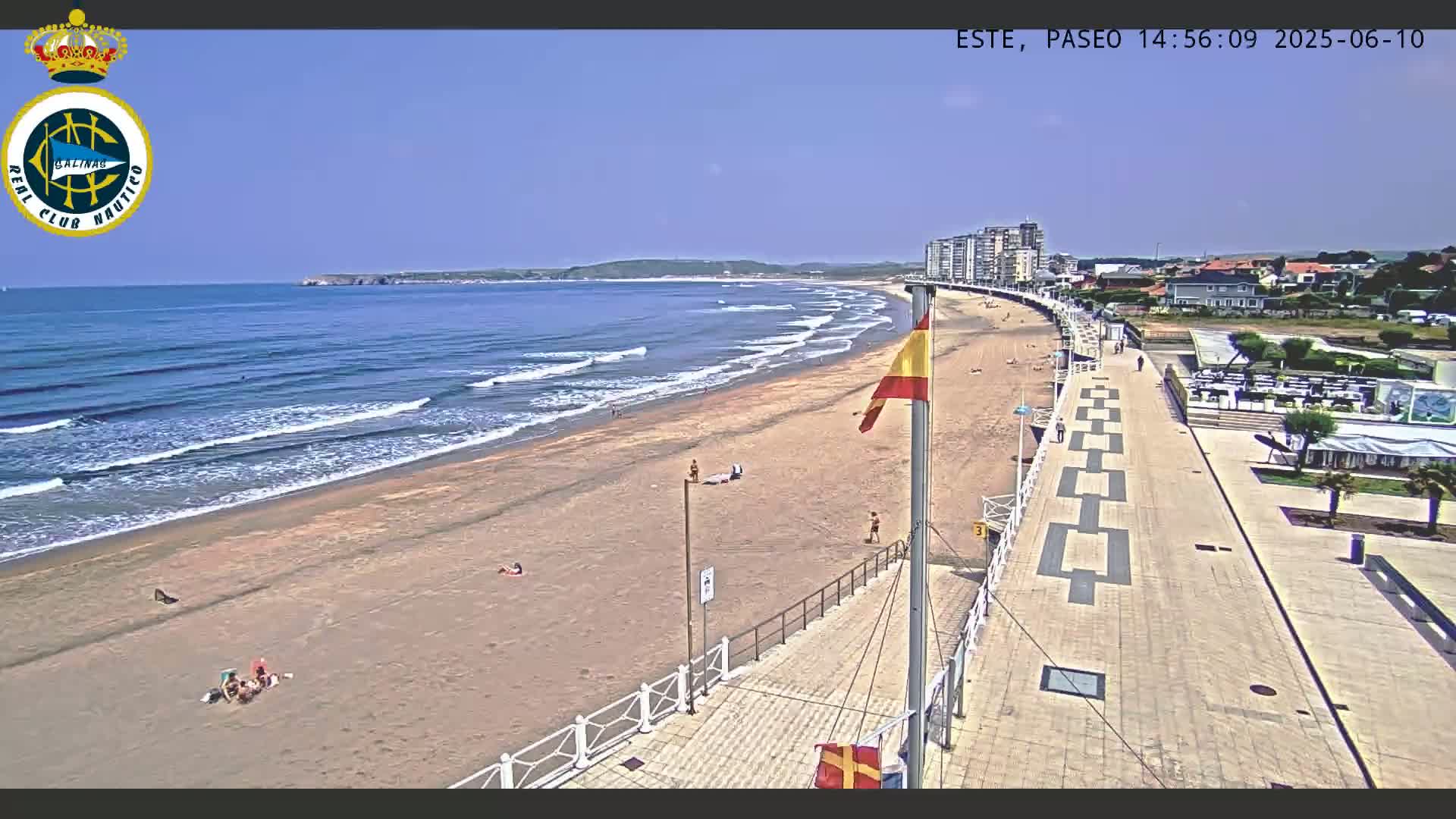 A sandy beach with gentle waves under a clear blue sky is bordered by a paved walkway leading to buildings, with a few people visible on the beach and walkway.