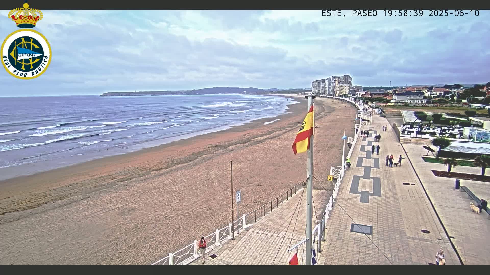 A sandy beach with gentle waves under a partly cloudy sky is bordered by a paved boardwalk with people walking, leading to buildings and restaurants.