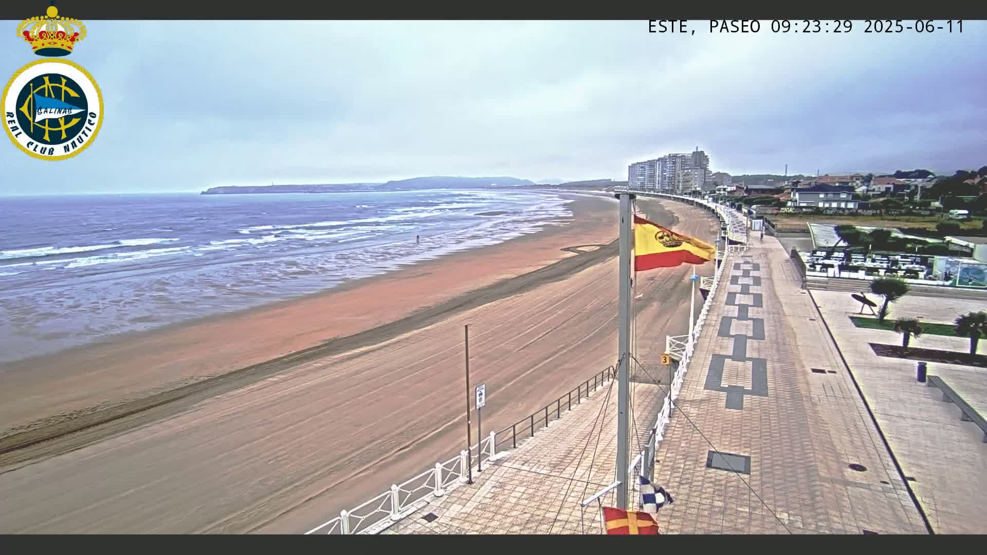 A sandy beach with gentle waves stretches along a paved promenade, under an overcast sky, beside buildings and a flagpole with a Spanish flag.