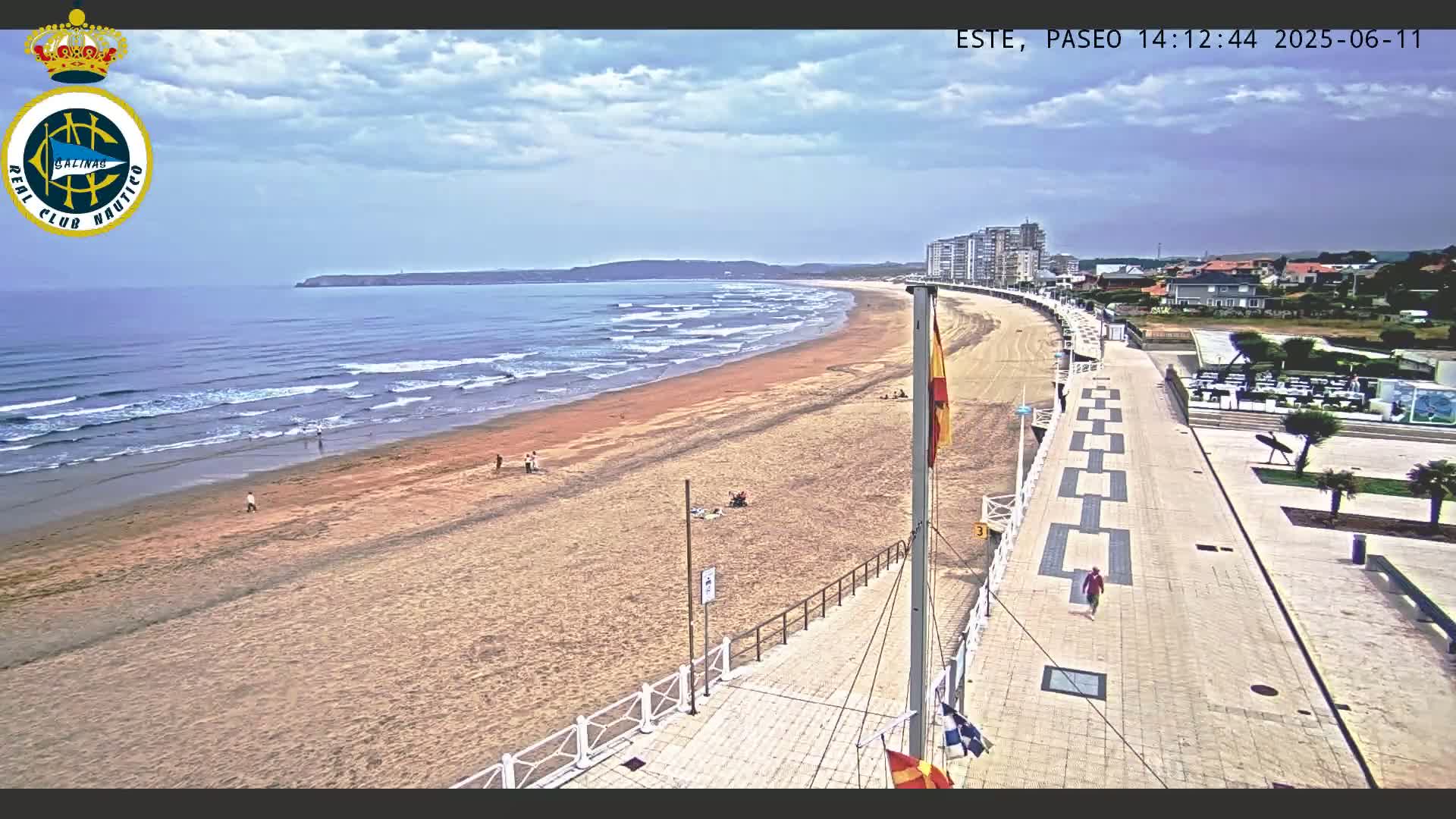 A sandy beach with gentle waves stretches along a coastline under a partly cloudy sky, next to a paved boardwalk lined with buildings.