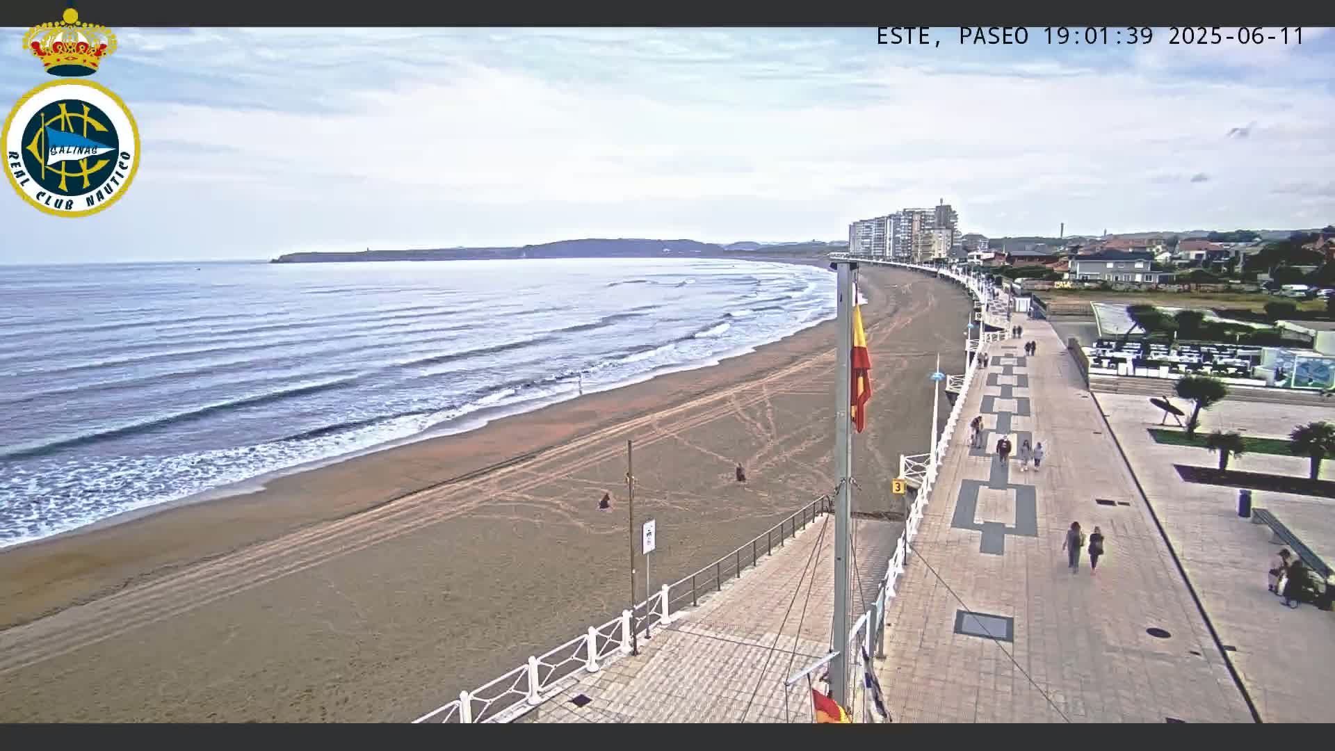 A sandy beach with gentle waves under a cloudy sky is bordered by a paved walkway with people strolling and buildings in the distance.