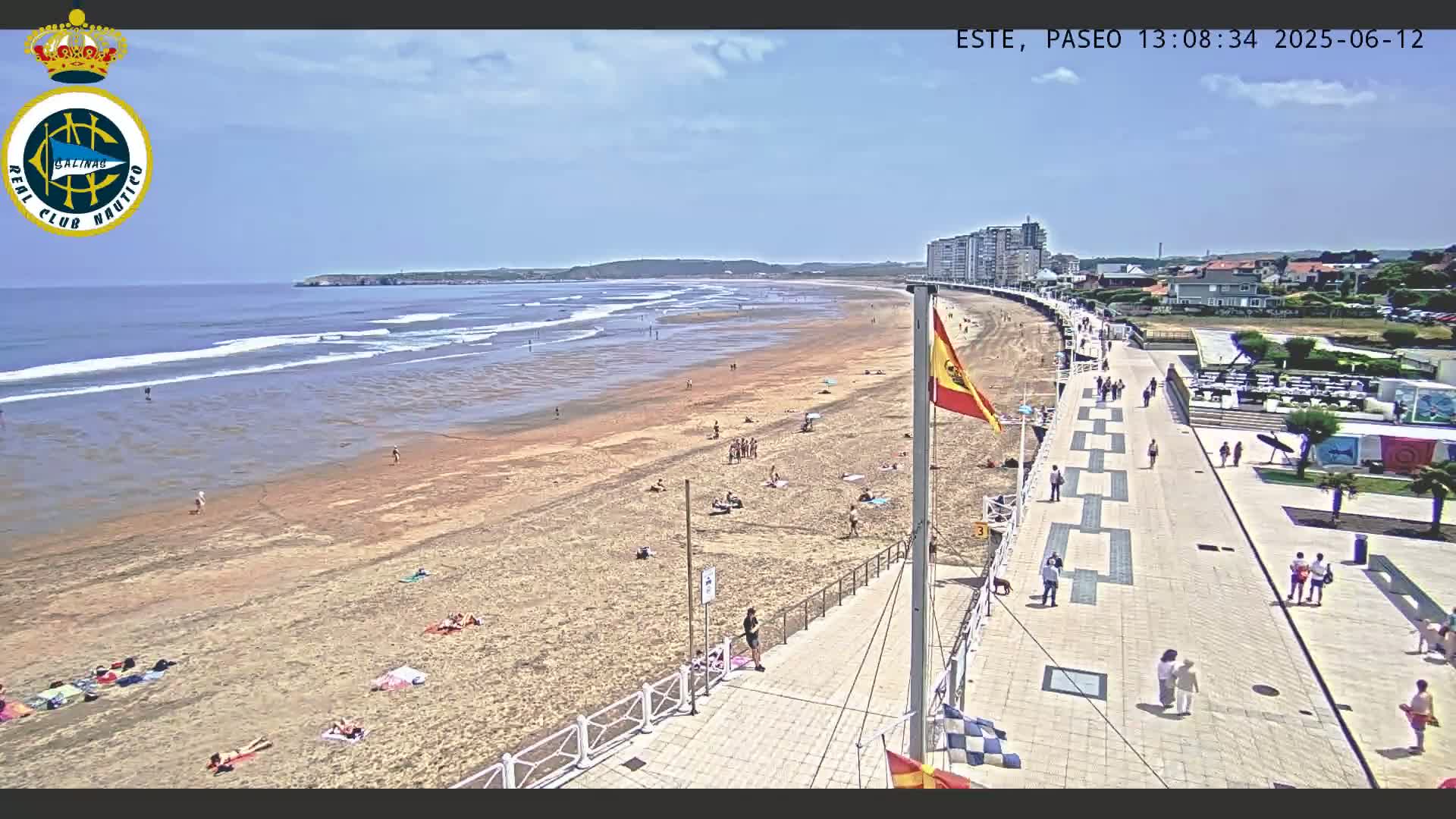 A sandy beach with people sunbathing and swimming, a boardwalk with pedestrians, and buildings in the background on a sunny day with some small waves.