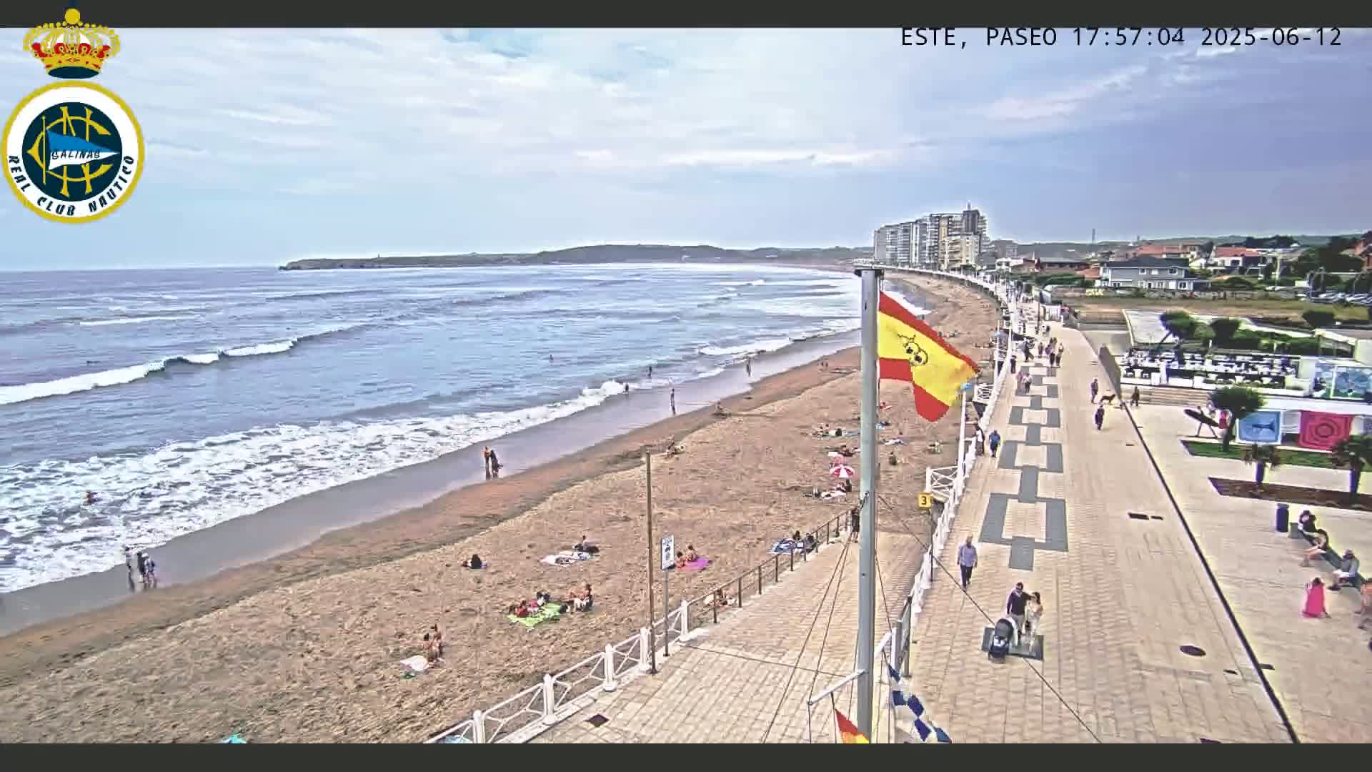 A sandy beach with moderate waves and scattered people under a partly cloudy sky is bordered by a paved promenade lined with buildings.