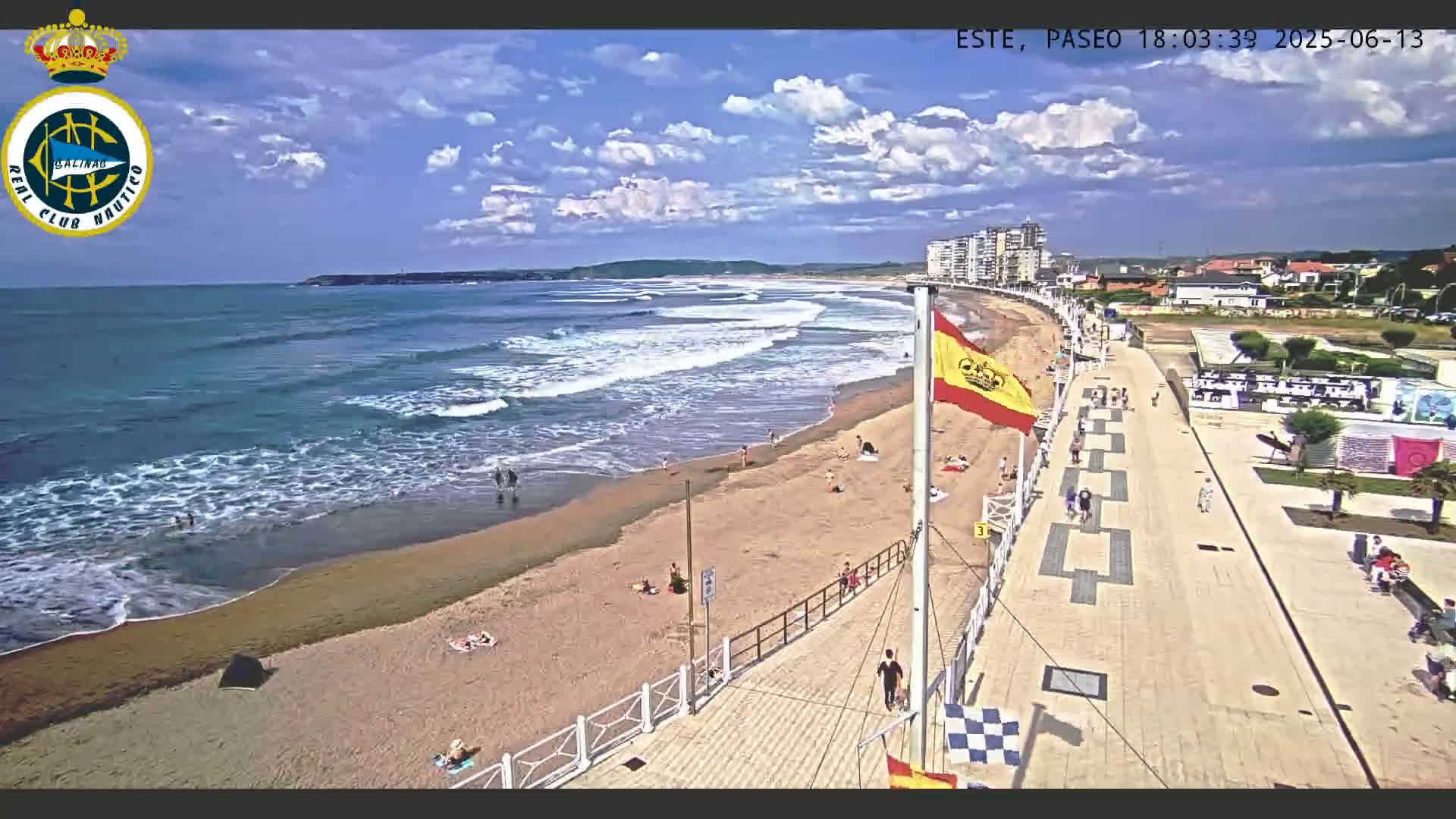 A sandy beach with moderate waves under a partly cloudy sky is shown, with a boardwalk and buildings visible in the background.