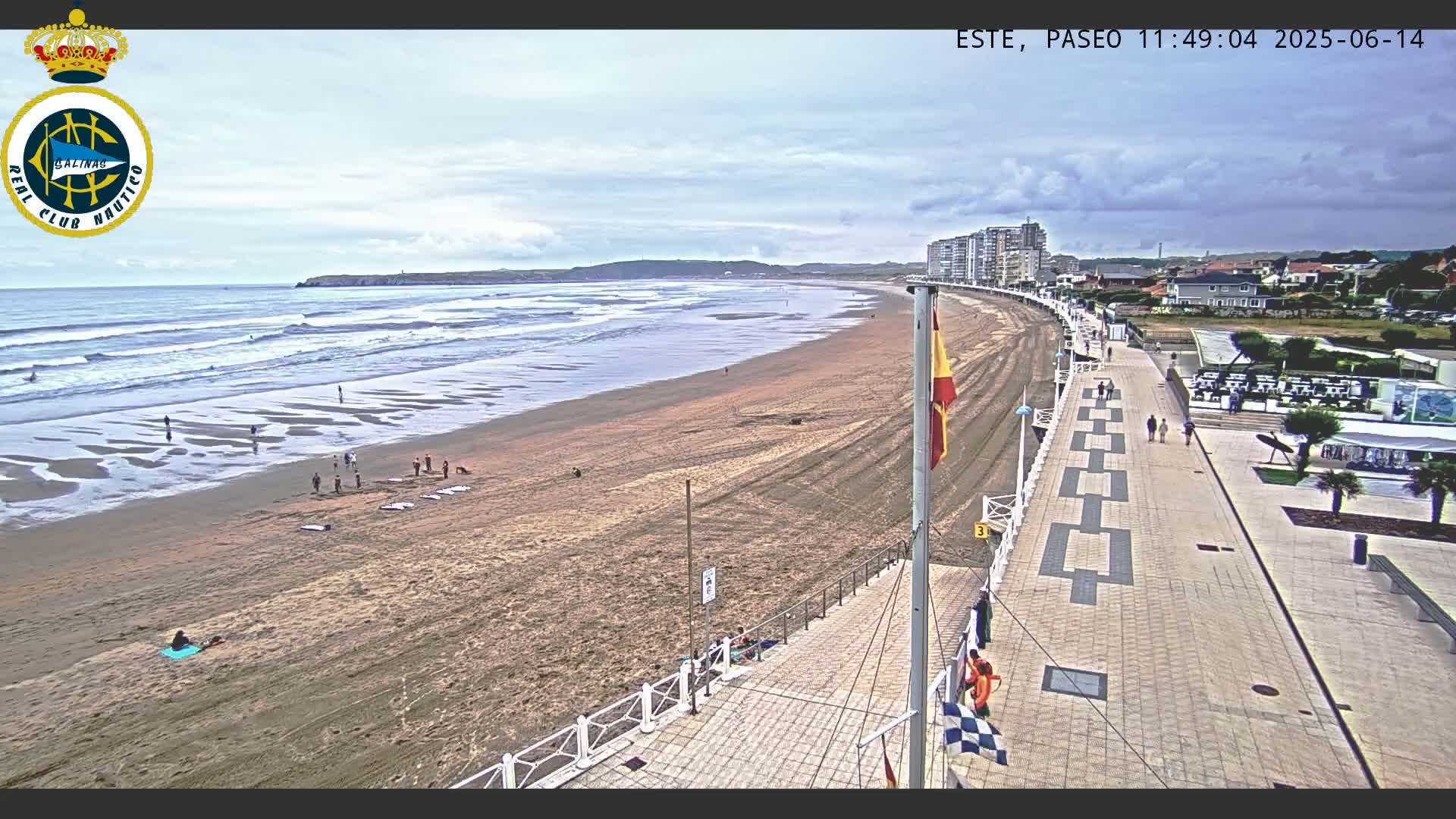 A sandy beach with moderate waves under a partly cloudy sky is bordered by a paved promenade with people walking and buildings in the background.