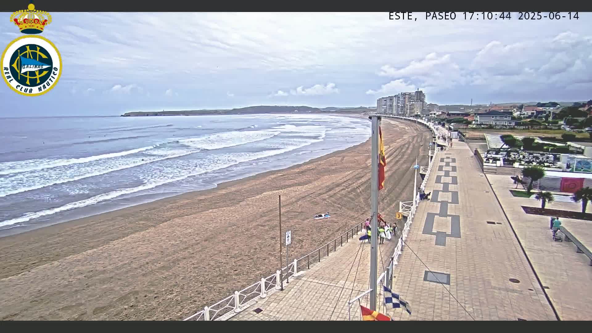 A sandy beach with gentle waves under a partly cloudy sky is bordered by a paved promenade and buildings.