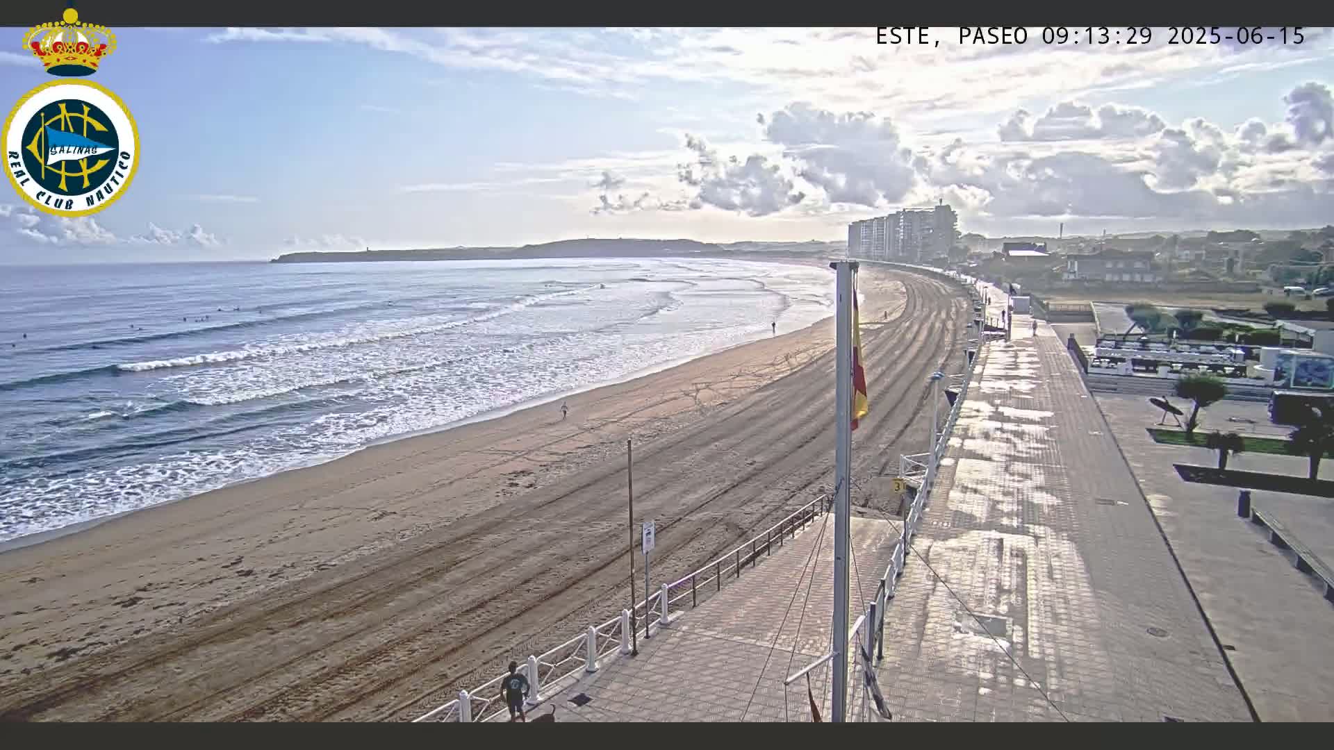 A sandy beach with gentle waves and a few surfers is bordered by a paved walkway and buildings under a partly cloudy sky.
