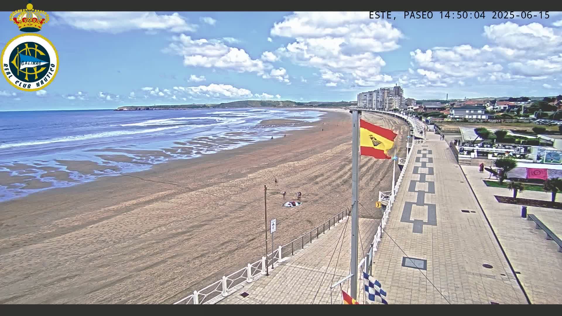 A sandy beach with small waves under a partly cloudy sky is next to a paved boardwalk with a Spanish flag and a few people visible.