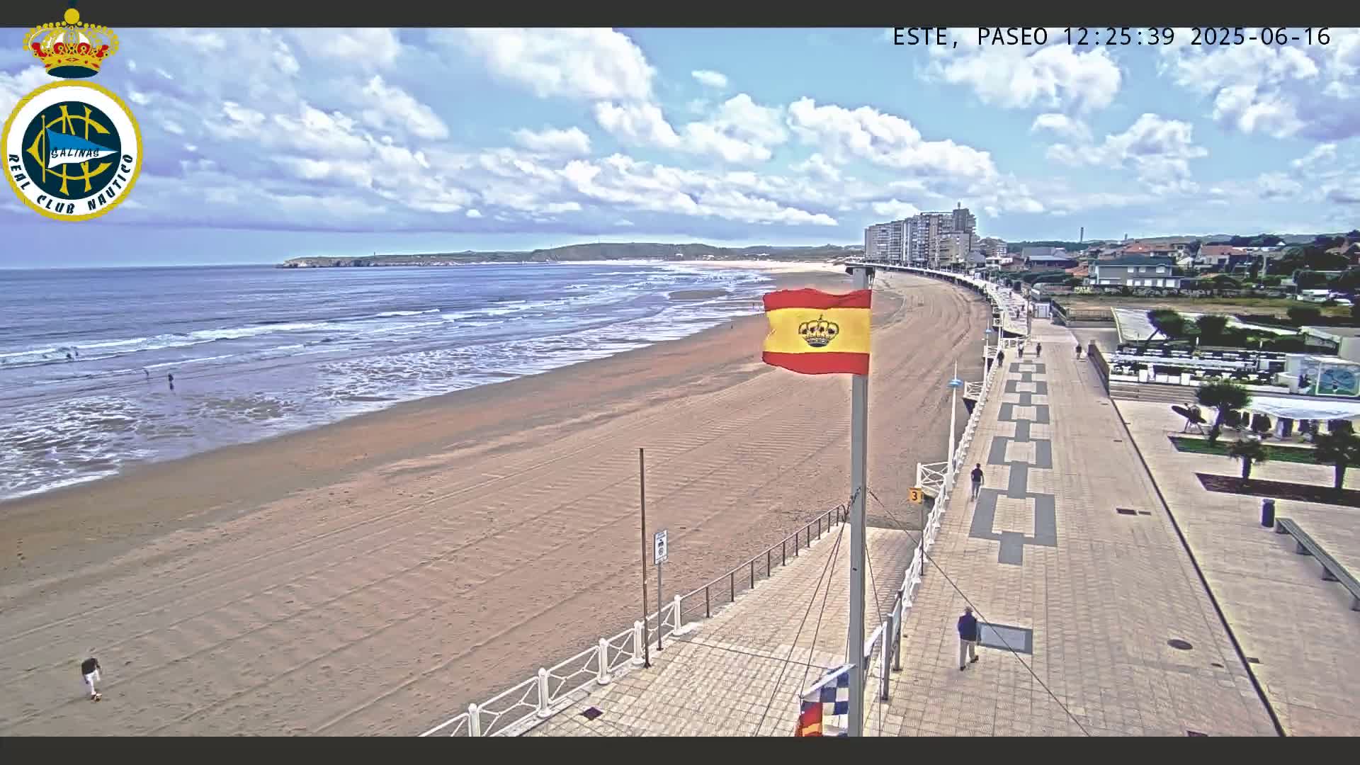 A sandy beach with moderate waves under a partly cloudy sky is adjacent to a paved walkway lined with buildings and a Spanish flag.