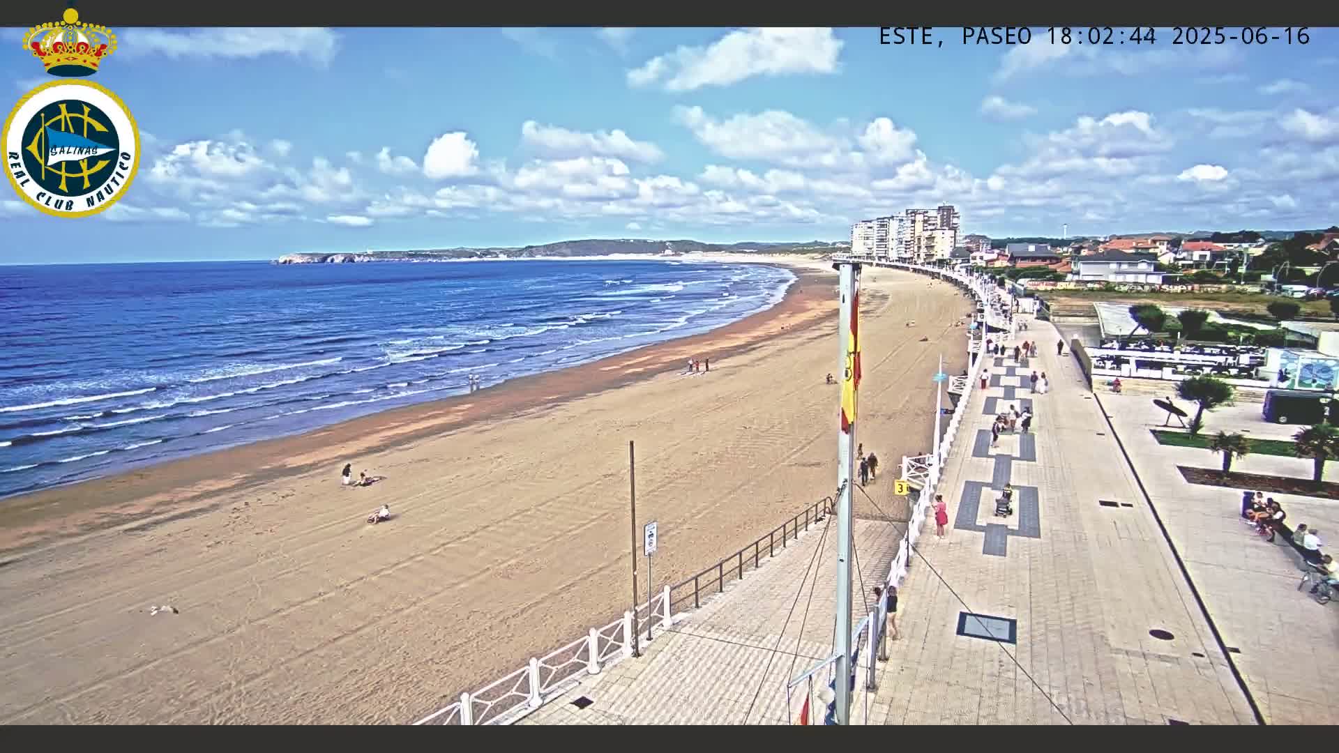 A sandy beach with gentle waves under a partly cloudy blue sky is bordered by a paved boardwalk with people walking along it and buildings visible in the distance.