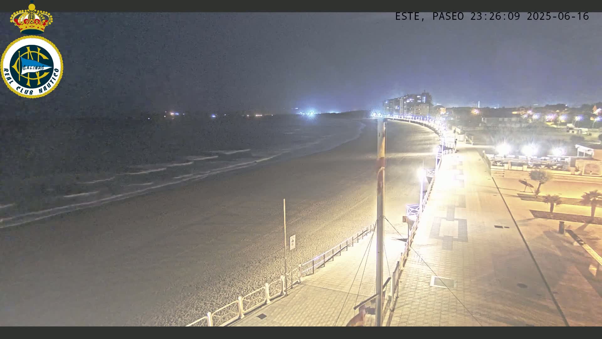 A nighttime view of a mostly empty beach with gentle waves, a paved boardwalk, and buildings in the distance under a dark sky.
