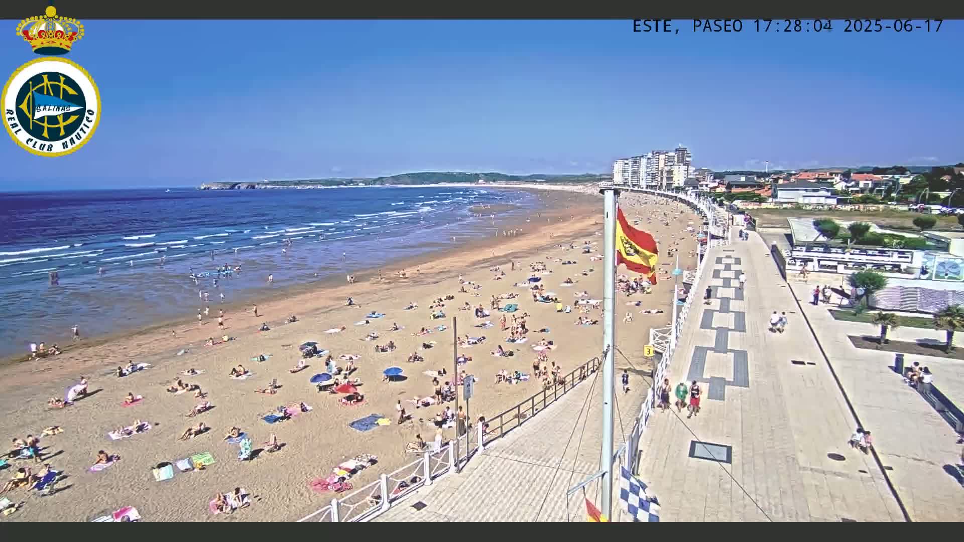 A sunny beach with many people sunbathing and swimming, next to a boardwalk and buildings, under a clear blue sky.
