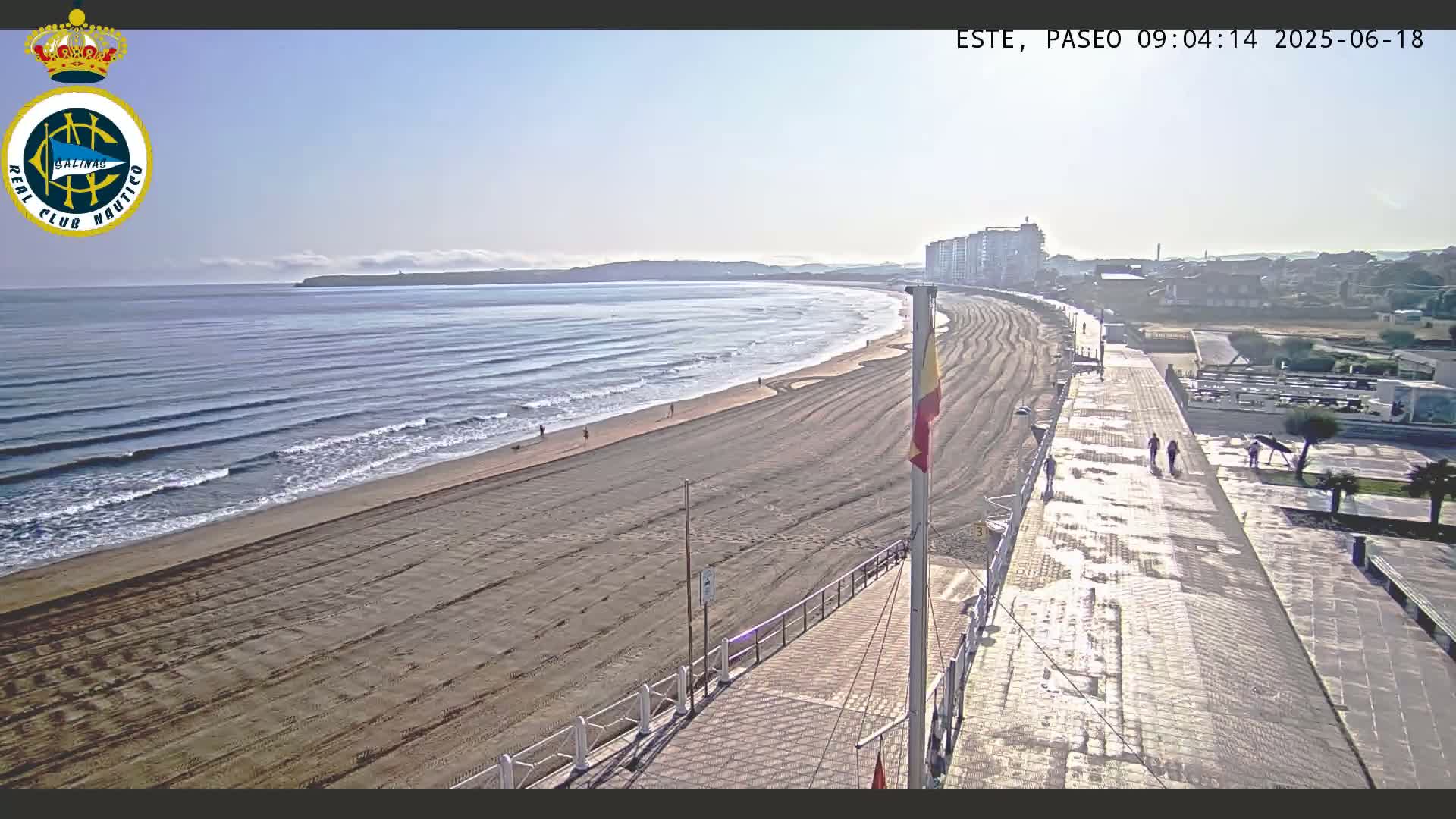 A mostly sunny day shows a long sandy beach with gentle waves, a boardwalk with a few pedestrians, and buildings in the distance.