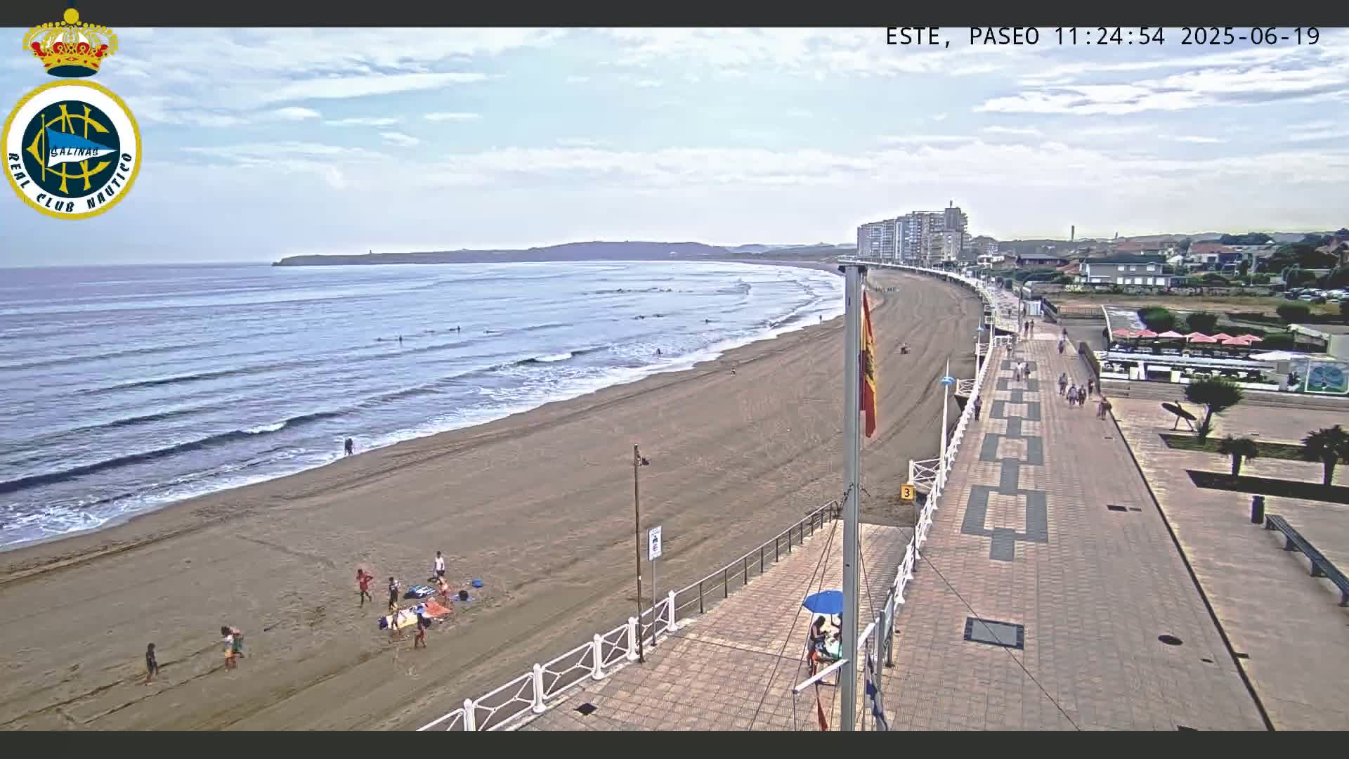 A sandy beach with people on it and a boardwalk leading to buildings is visible under a partly cloudy sky.