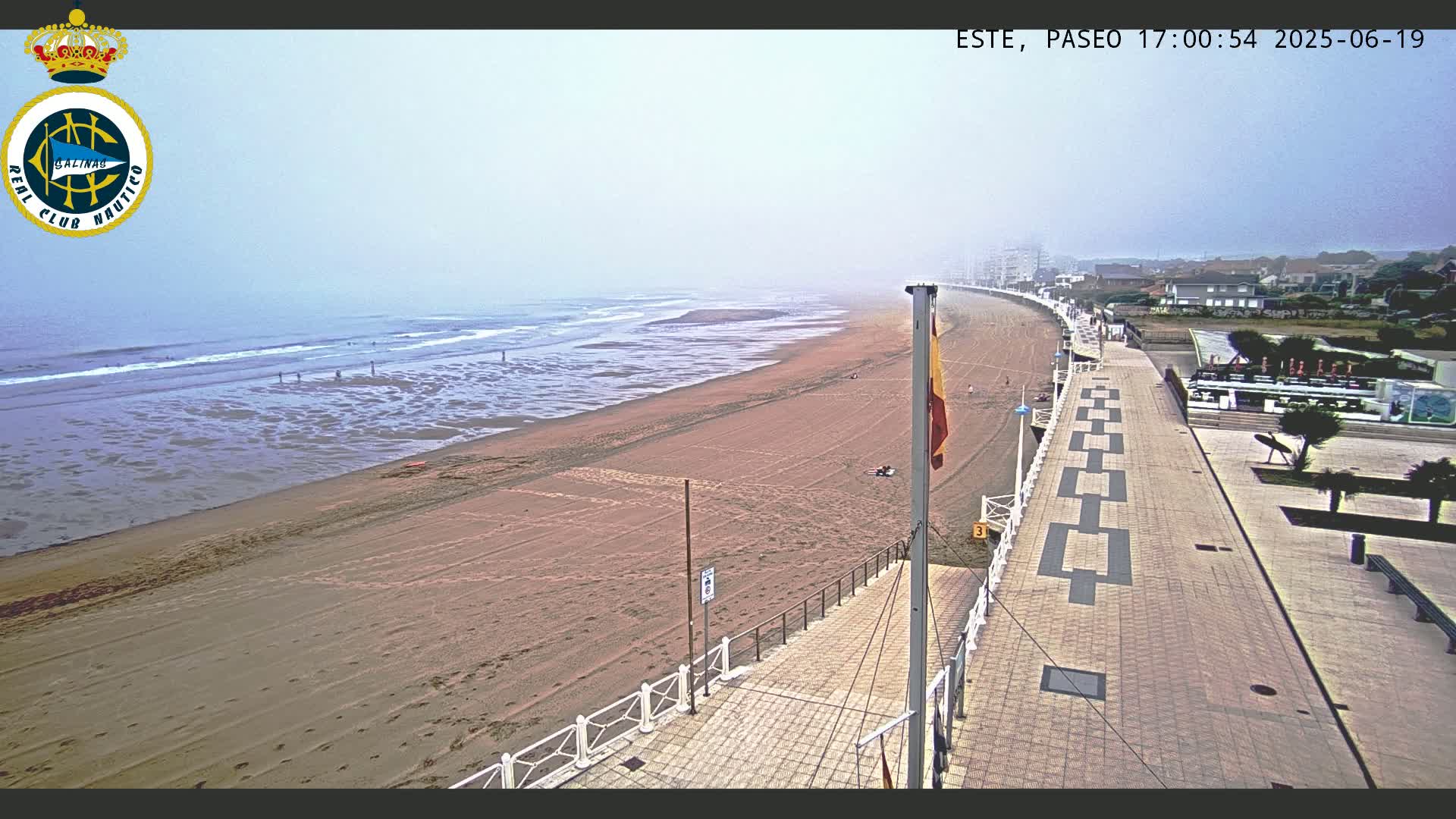 A sandy beach stretches along a paved boardwalk on a foggy day, with a flagpole and some people visible in the distance.
