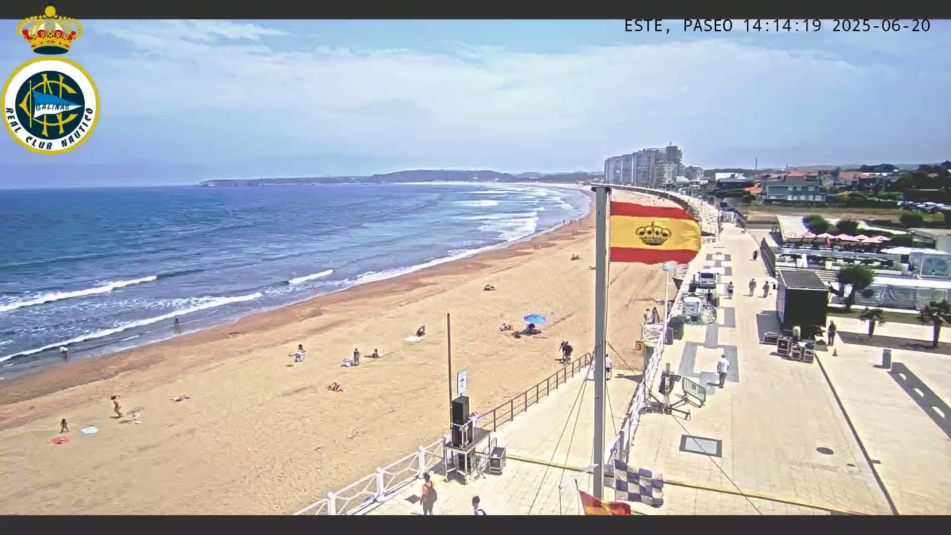 A sandy beach with a few people sunbathing and swimming is bordered by a paved walkway leading to buildings, all under a partly cloudy sky.