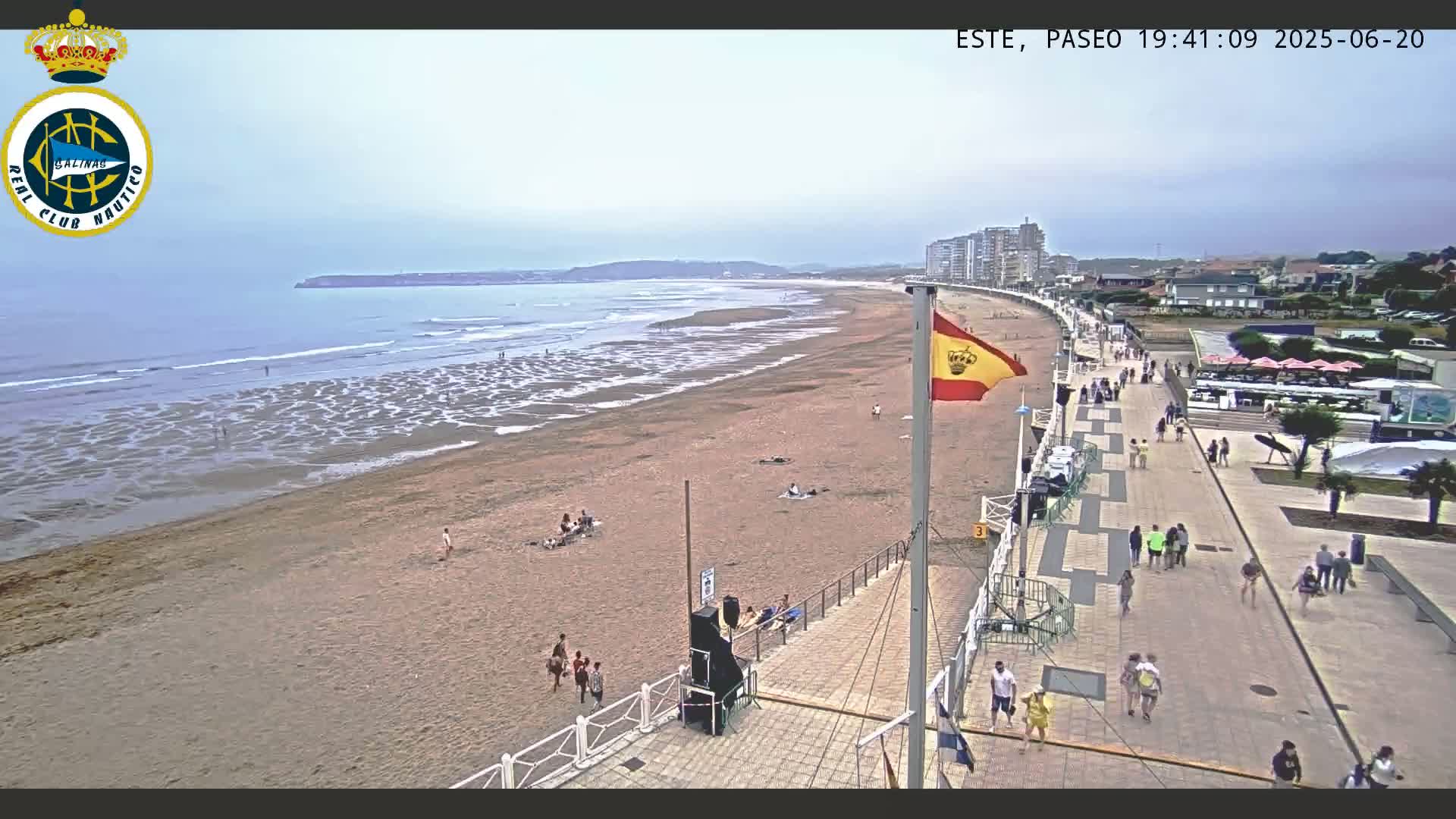 A sandy beach with scattered people stretches alongside a paved boardwalk, with buildings and a Spanish flag visible in the background under an overcast sky.