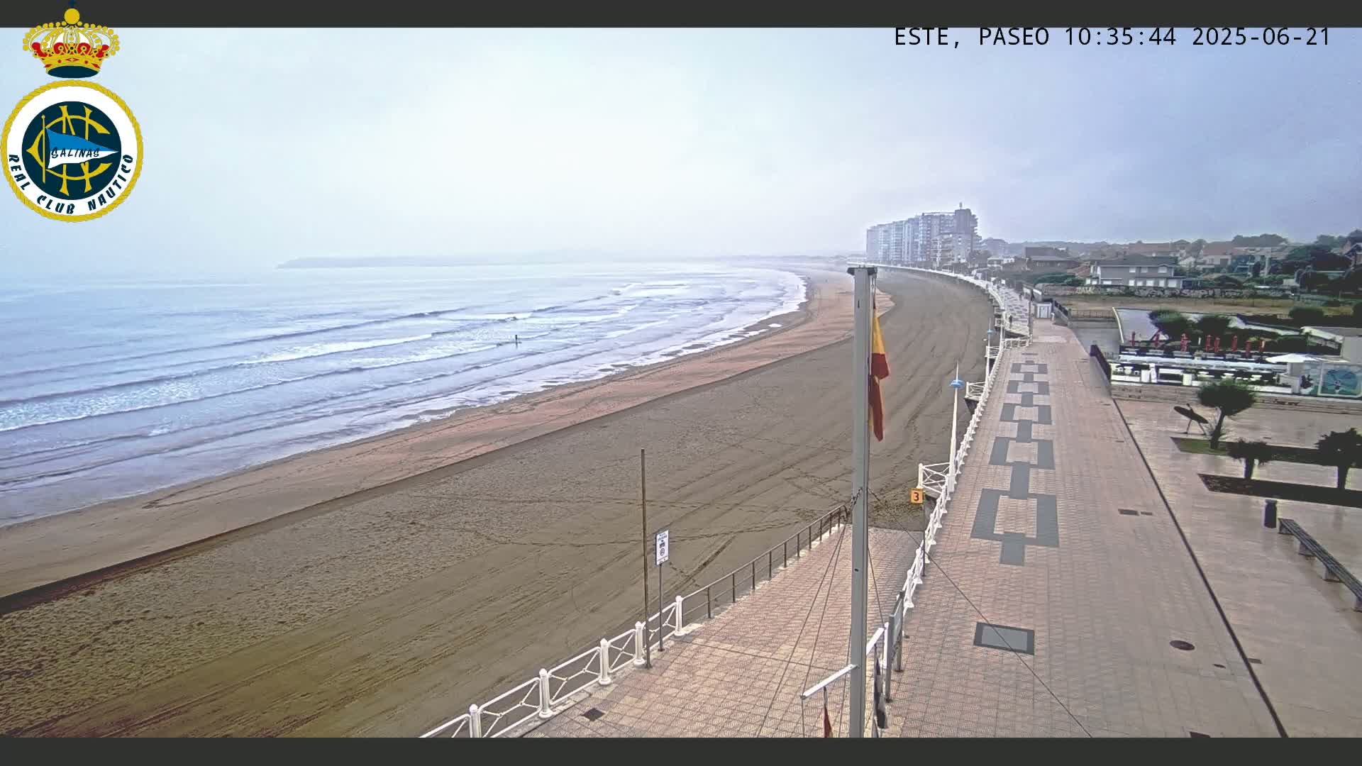 A sandy beach with gentle waves under an overcast sky is seen from above, adjacent to a paved walkway and buildings.