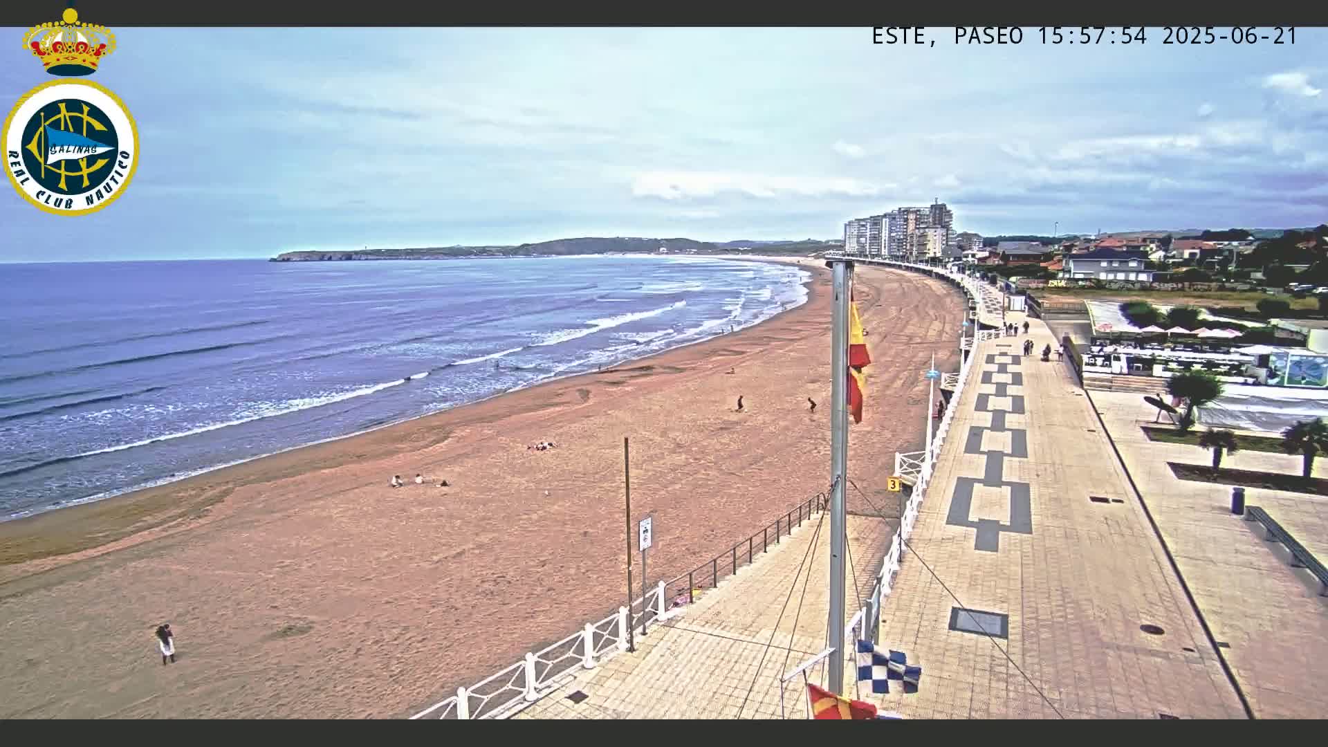 A sandy beach with gentle waves under a partly cloudy sky is overlooked by a paved boardwalk lined with buildings.
