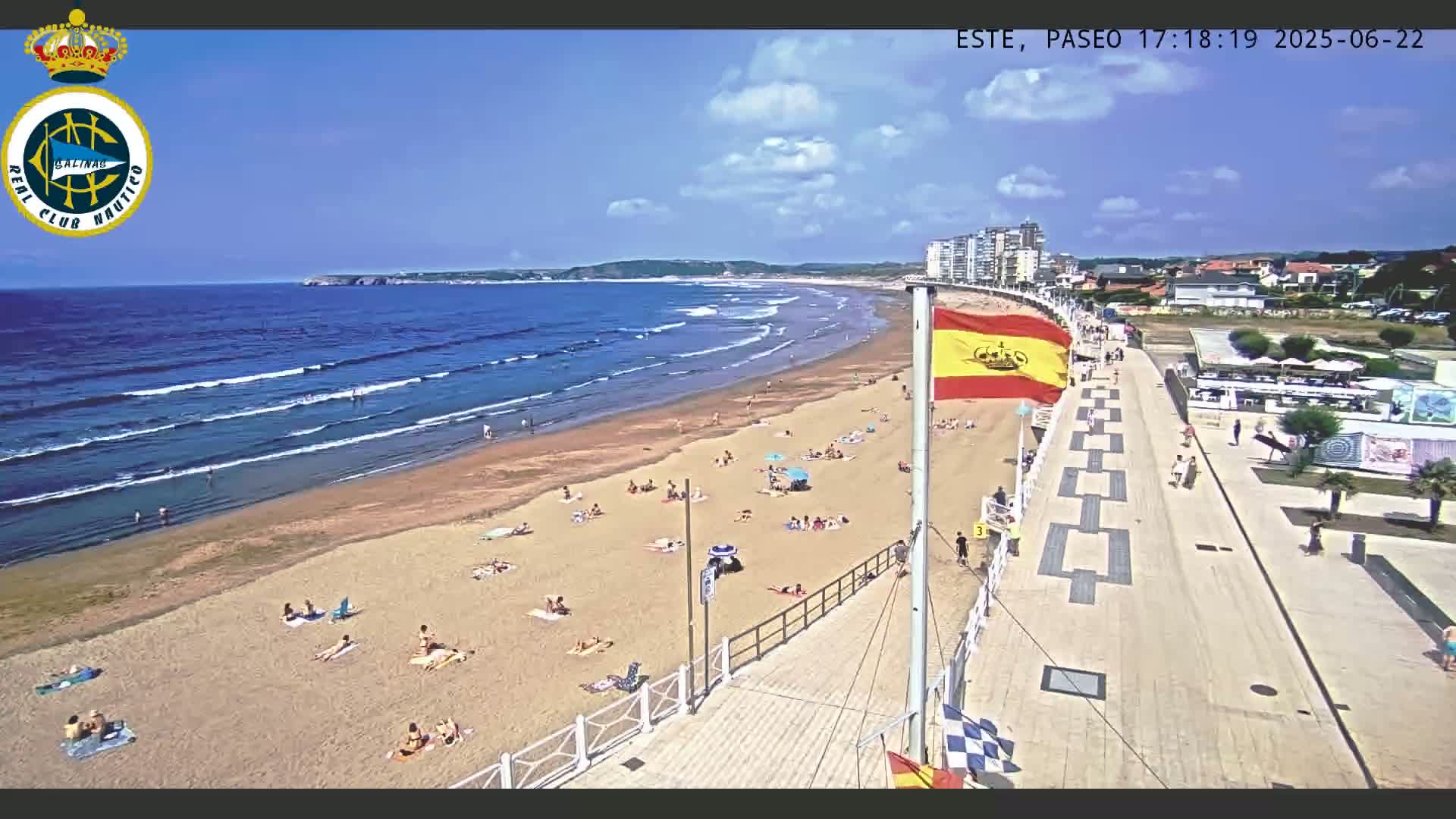 A sandy beach with many people sunbathing and swimming, bordered by a paved walkway and buildings, under a partly cloudy sky.