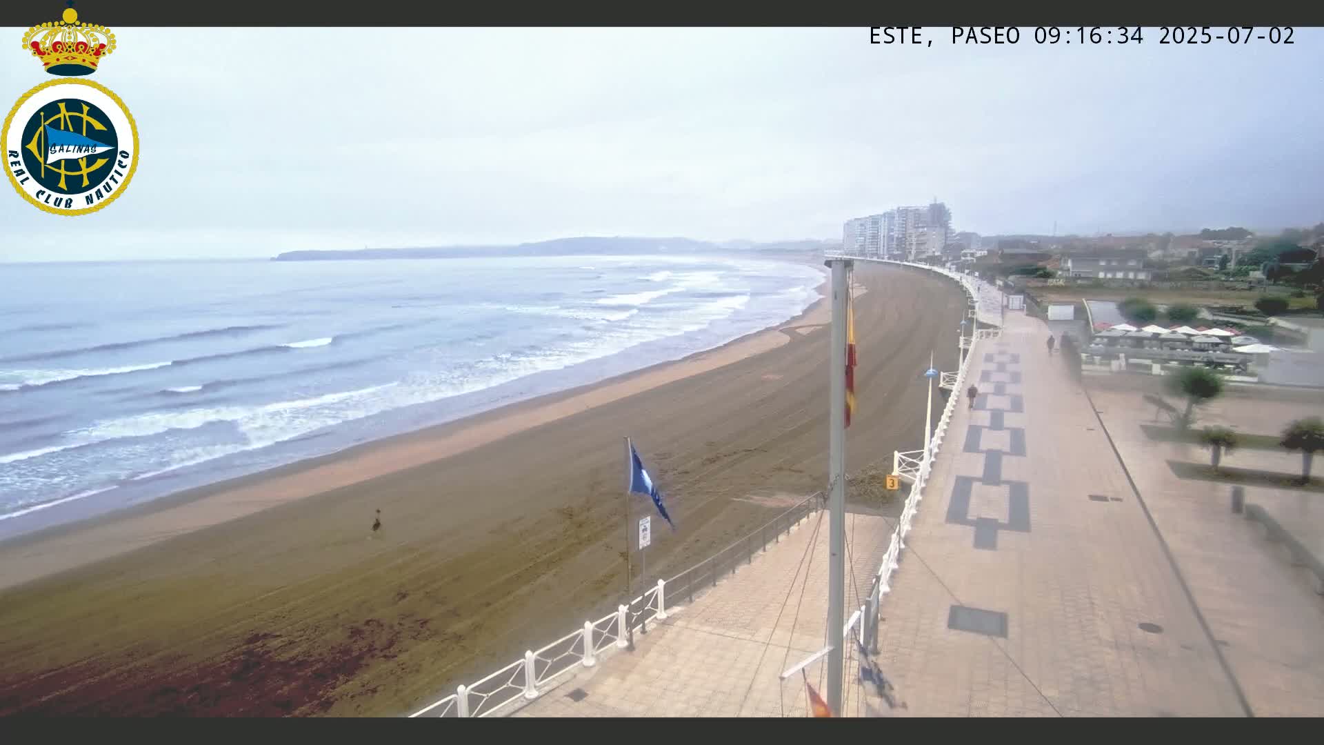 A sandy beach with moderate waves under an overcast sky is bordered by a paved walkway leading to buildings.