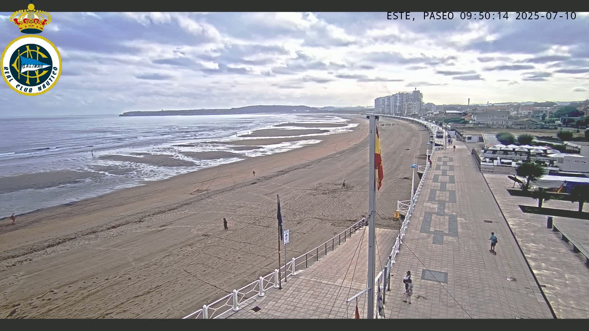 A sandy beach with gentle waves under a partly cloudy sky is bordered by a paved walkway and buildings.
